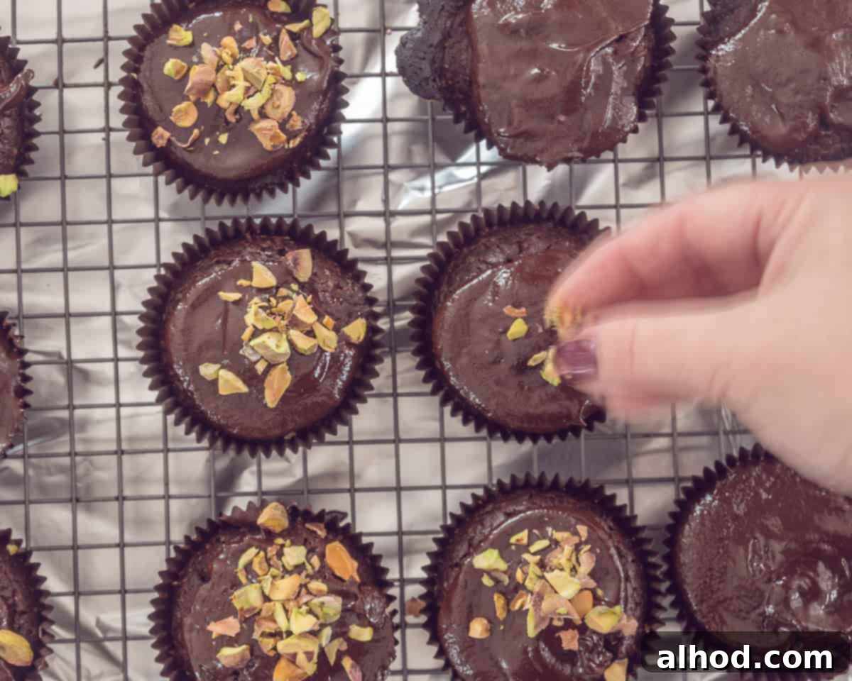 Decadent Triple Chocolate Cupcakes 7 A close-up shot of ganache-covered chocolate cupcakes on a cooling rack, with a hand delicately sprinkling chopped salted pistachios over them, adding a perfect crunch.