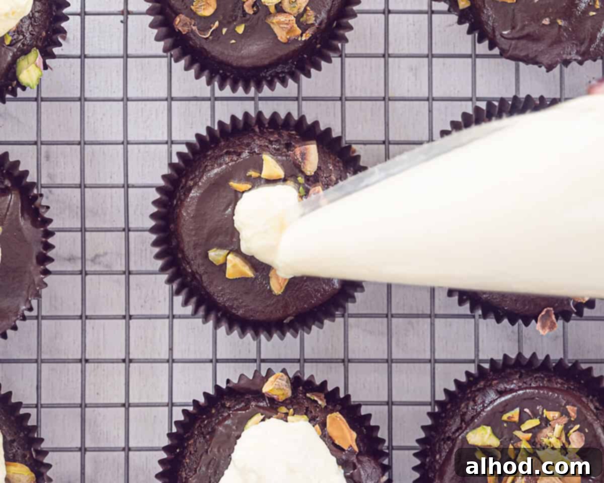 Decadent Triple Chocolate Cupcakes 8 A piping bag meticulously creating a swirl of creamy white chocolate buttercream on top of a chocolate cupcake, which is already coated with ganache and sprinkled with pistachios.