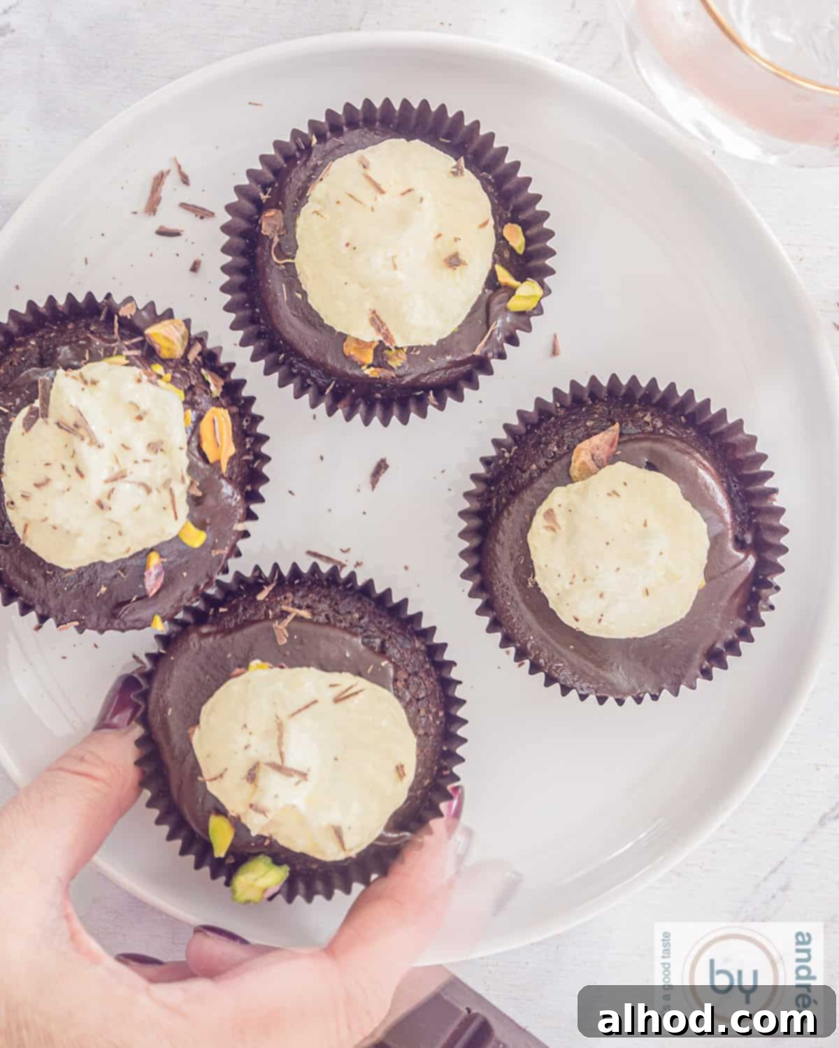 Decadent Triple Chocolate Cupcakes 9 A stunning top-down view of four Triple Chocolate Cupcakes with distinct layers of chocolate cake, ganache, white chocolate buttercream, and a sprinkle of pistachios, with a hand reaching for one.