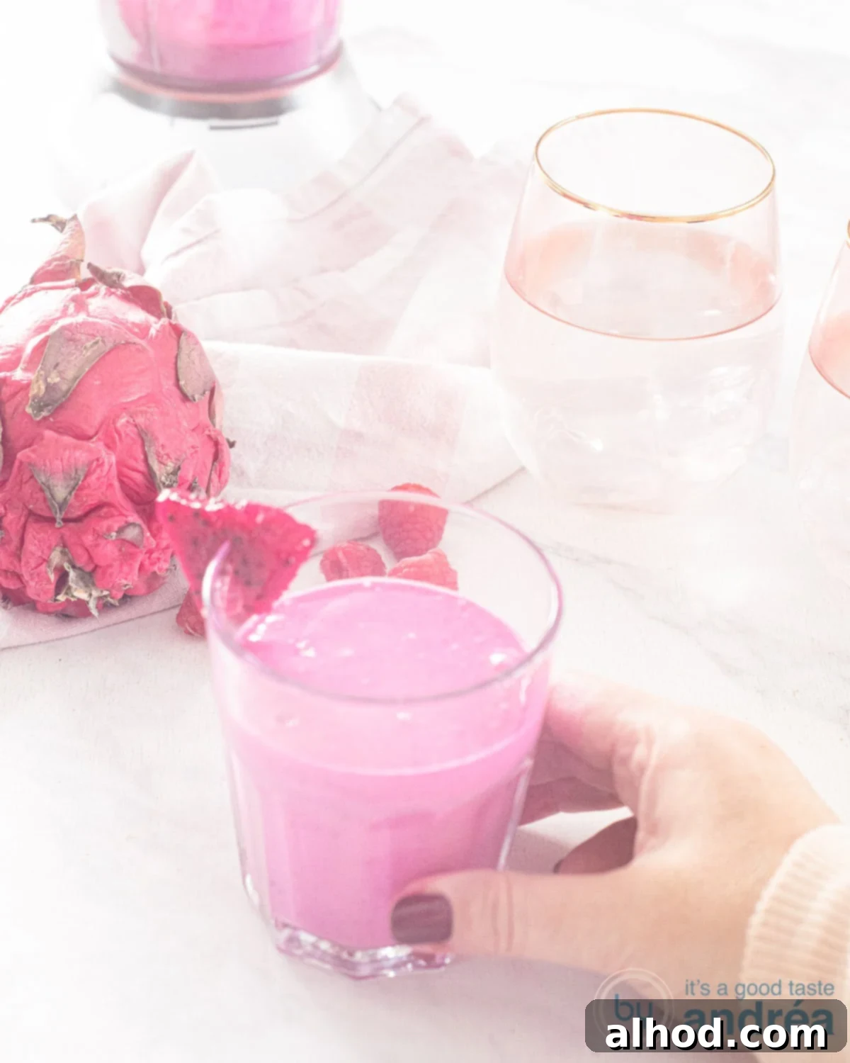 Tropical Dragon Blend 2 A glass filled with dragon fruit smoothie on a white surface. A hand grabs the glass. In the white background, glasses and pitahaya fruit.
