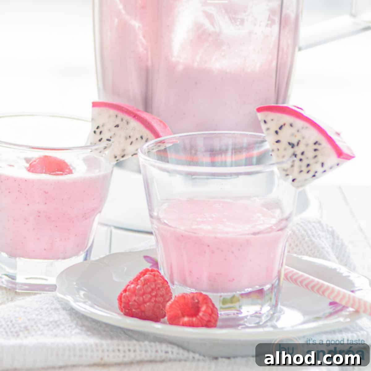 Tropical Dragon Blend 4 A square photo with a white plate on top of which a glass filled with dragon fruit smoothie with raspberries and a slice of dragon fruit. A white background