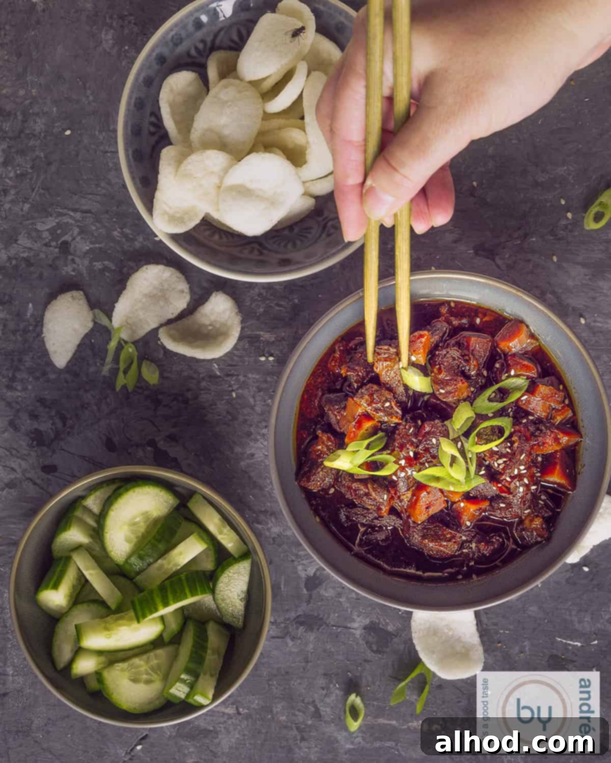 Easy Crockpot Mongolian Beef 5 A photo with three grey bowls filled with mongolian beef, prawn crackers and cucumber salad. Two chopsticks taking some meat