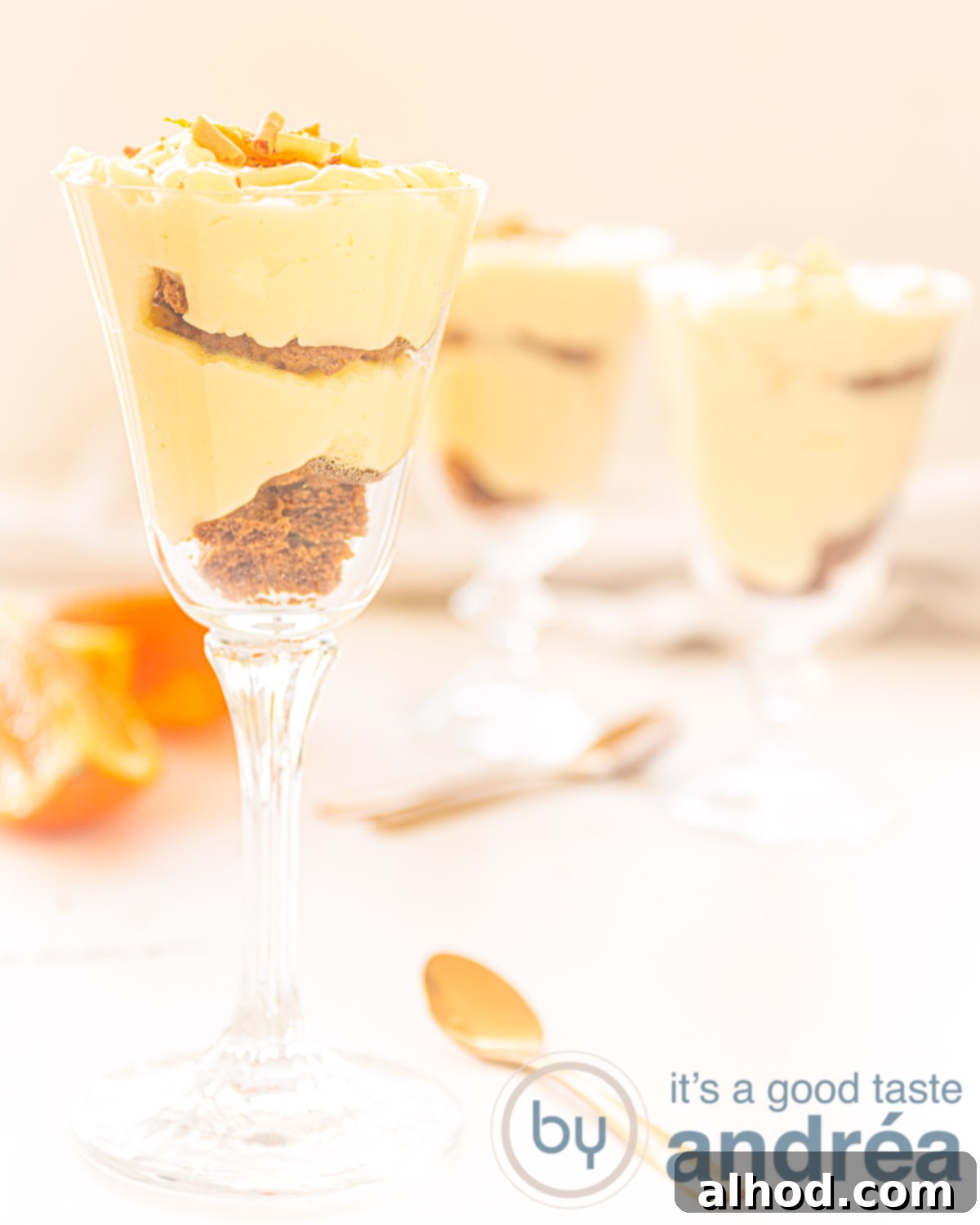 Zesty Orange Delight 4 Three elegant glasses filled with layers of orange custard and biscuits, set against a pristine white background adorned with fresh oranges.