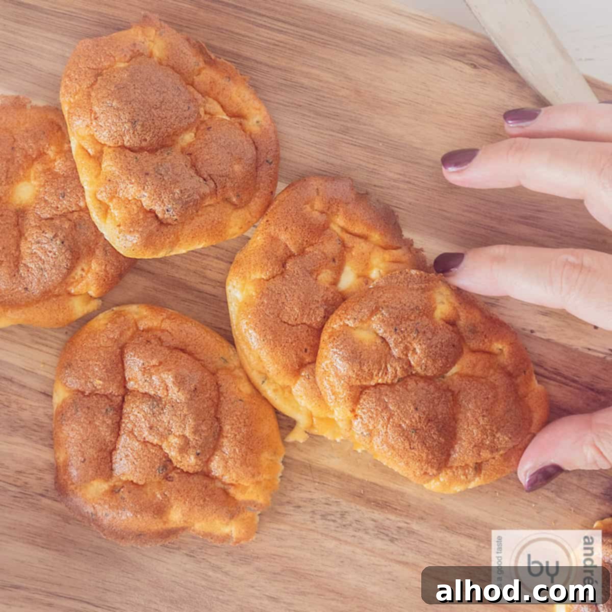 Cottage Cheese Cloud Bread 3 A square photo with a wooden board with cloud breads on it. A hand grabs an oopsie