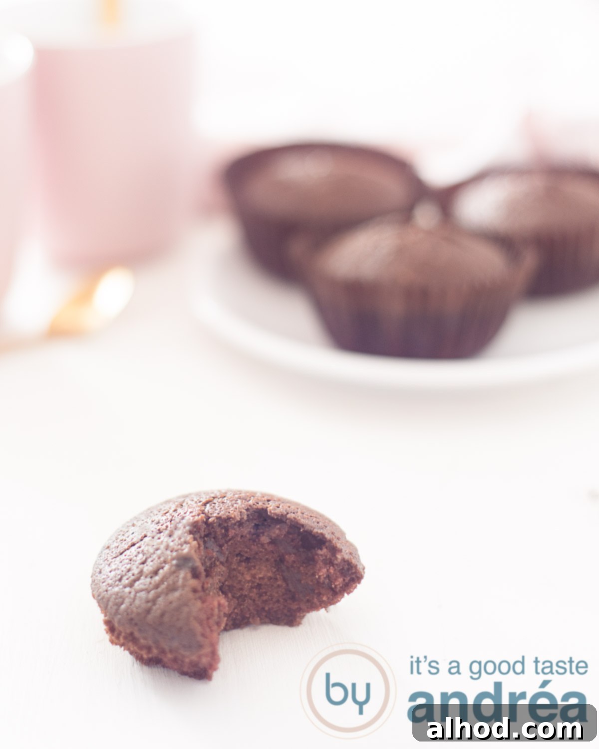 A close-up shot of a single dairy-free chocolate cupcake in the foreground, with a delicious bite already taken, revealing its perfectly moist and fluffy interior. In the soft-focus background, a white plate holds three more beautifully baked chocolate cupcakes next to a cozy pink mug, inviting a moment of sweet indulgence.