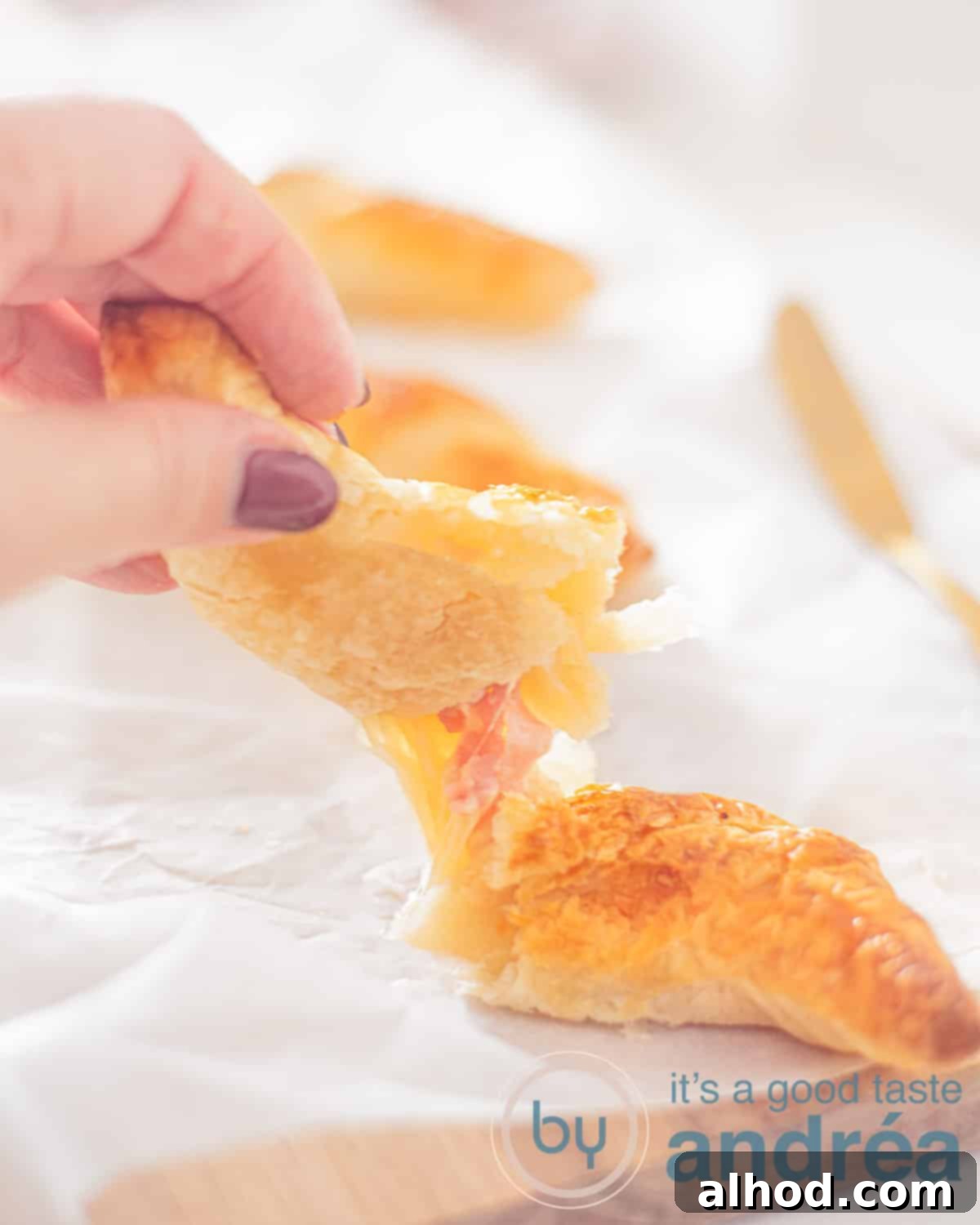 Flaky Ham and Cheese Puff Pastry Croissants 2 An elevation view of a croissant filled with ham and cheese, cut open and held up, revealing threads of melted cheese and ham. A perfect golden crust surrounds a savory interior.