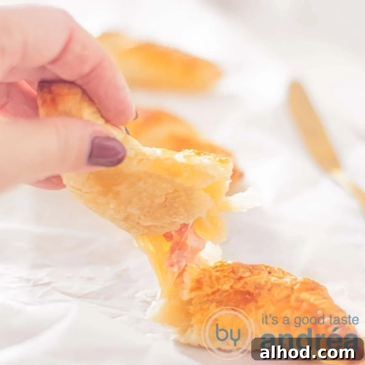 Flaky Ham and Cheese Puff Pastry Croissants 6 A square photo with three ham and cheese croissants in a row. The first croissant is broken open and a hand is holding a piece. In between you see melted cheese and ham.