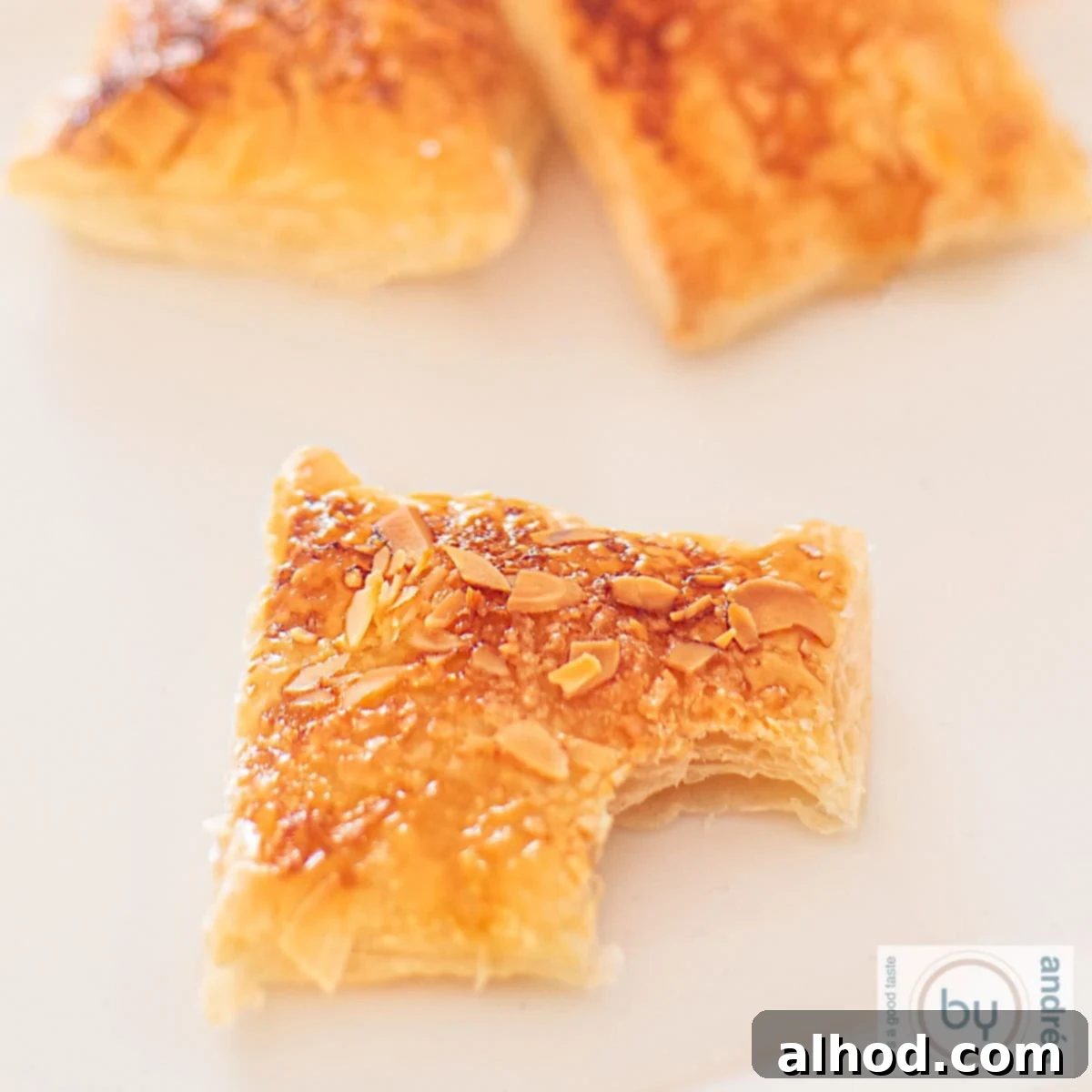 Flaky Zesty Lemon Puff Pastry Cookies 4 A square photo of lemon cookies with an almond layer. A bite has been taken from the front cookie.