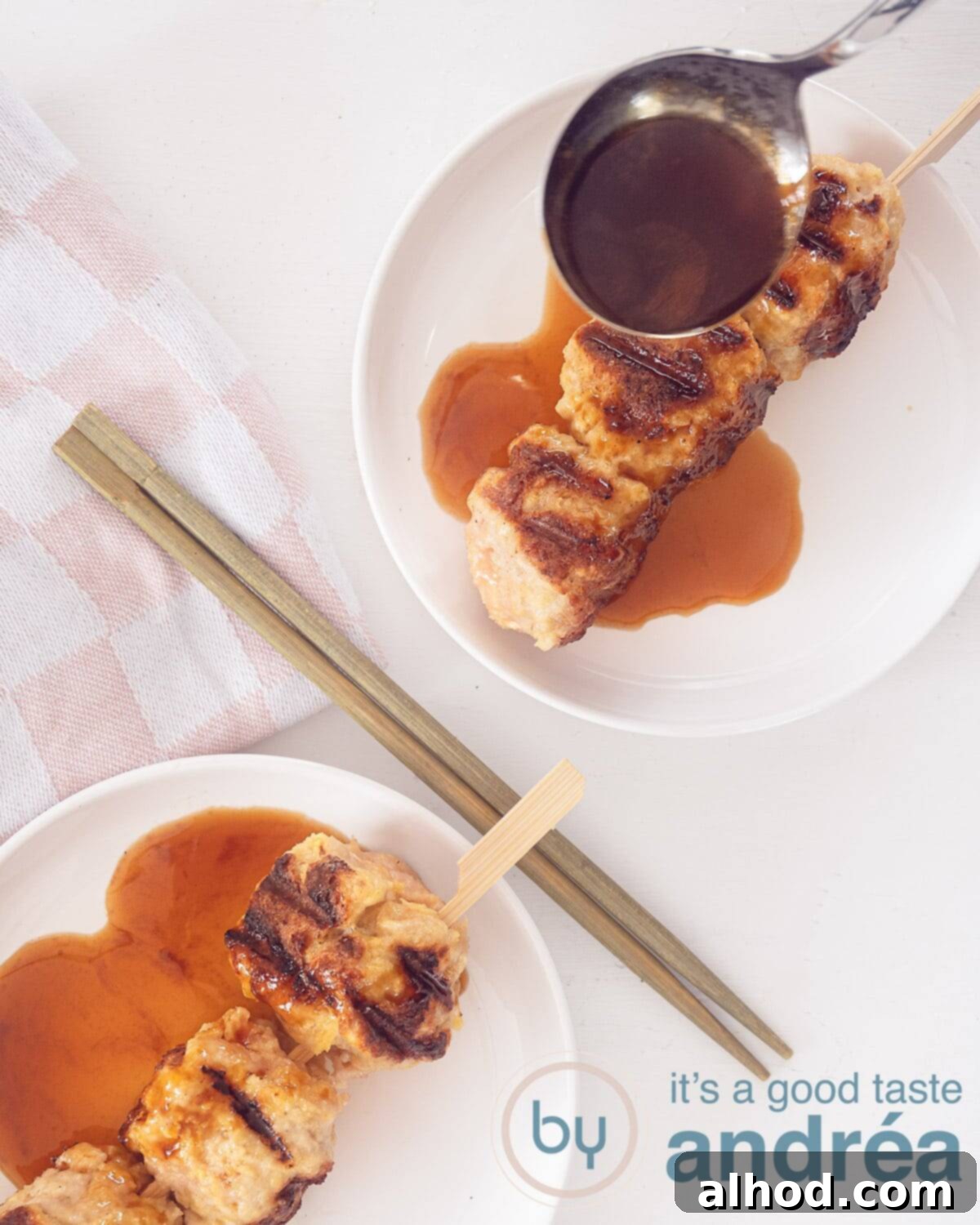 Tsukune Juicy Japanese Chicken Meatball Skewers with Teriyaki Sauce 3 A photo from above of two ground chicken kebabs with sauce on a white plate on a white background. Two golden chopsticks in between.
