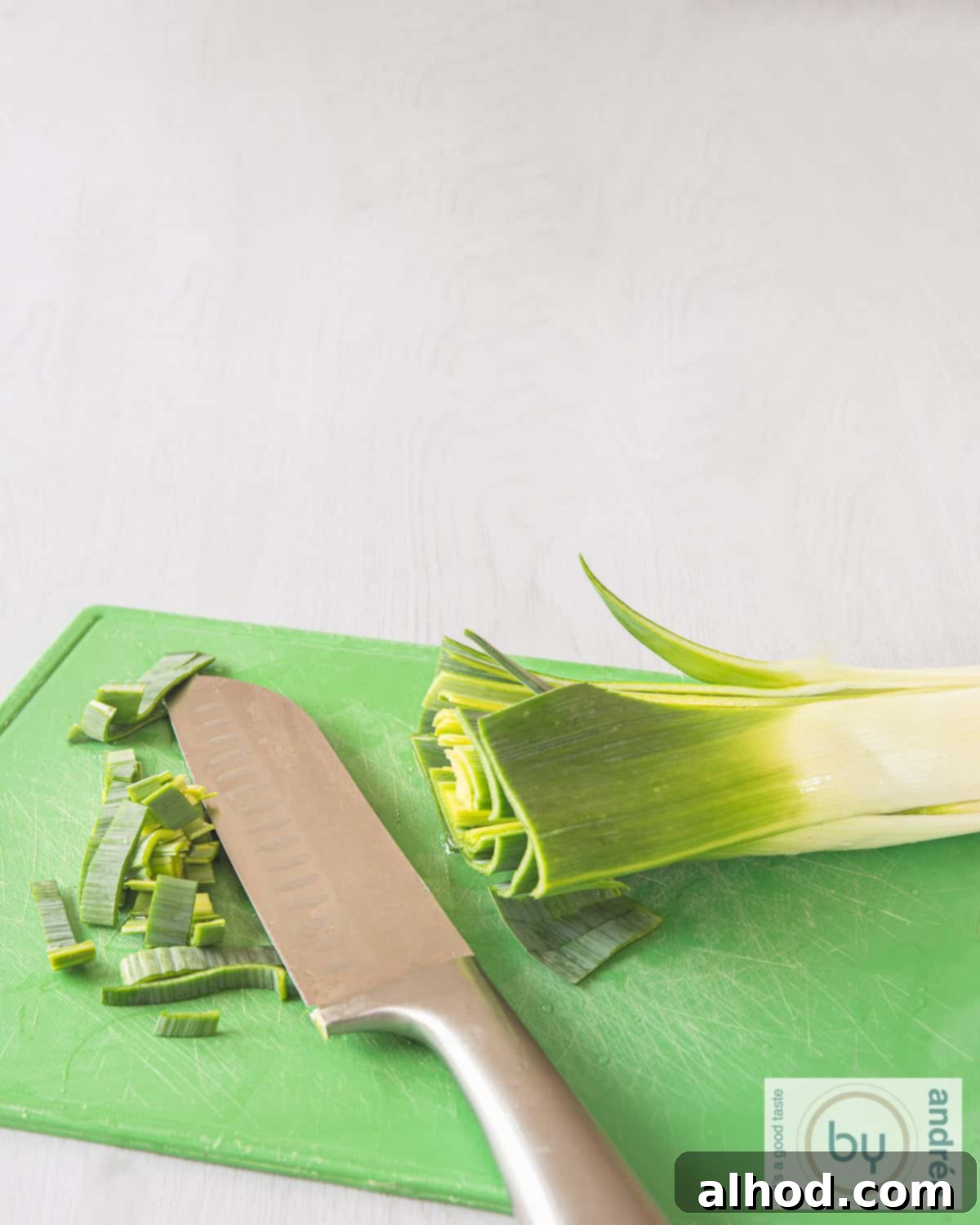 An aerial photo with a green cutting board on which a leek lies. A knife cuts strips from it.