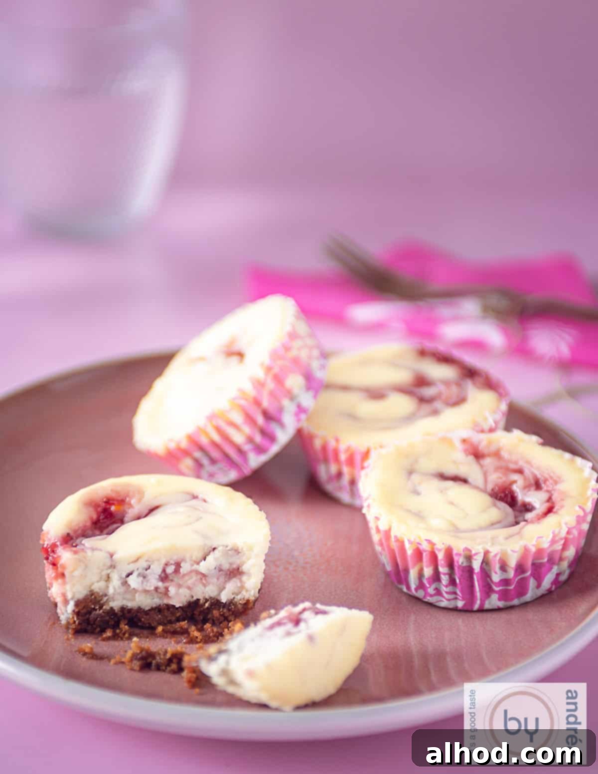 An aerial photo of a pink background with a pink plate with four marble cheesecake cupcakes with strawberries. 1 cupcake broken in half, so that the content is clearly visible.
