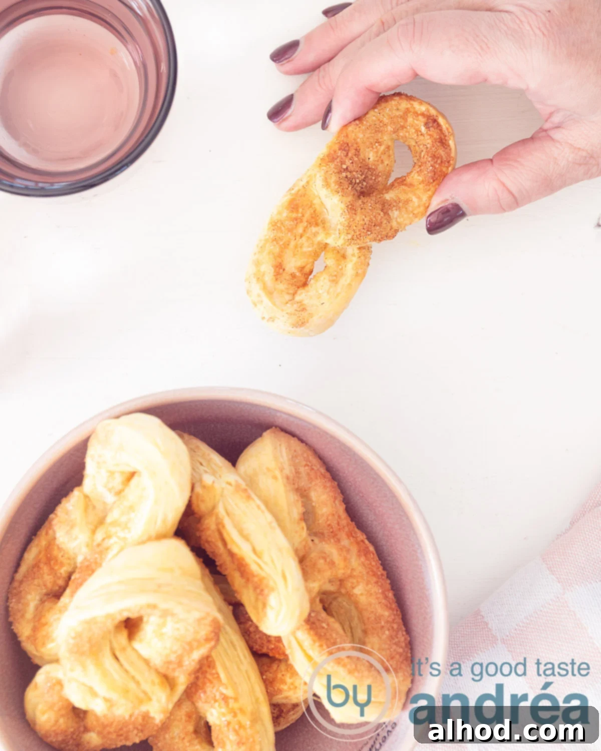 Sweet pretzels, also known as Krakelingen, neatly arranged in a pink bowl. A hand reaches into the upper left corner, gracefully taking one of the delicious cookies. The scene is set against a clean, inviting white background.