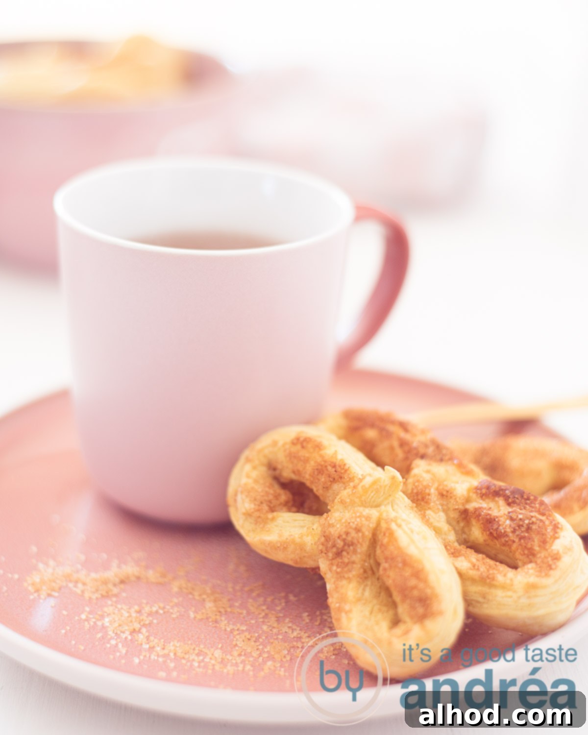 Krakelingen Sweet Dutch Pretzel Delights 5 Three golden-brown Krakelingen, Dutch sweet pretzel cookies, presented elegantly on a pink plate, accompanied by a pink mug filled with steaming tea. In the soft, blurred white background, a pink bowl brimming with more cookies hints at abundance.