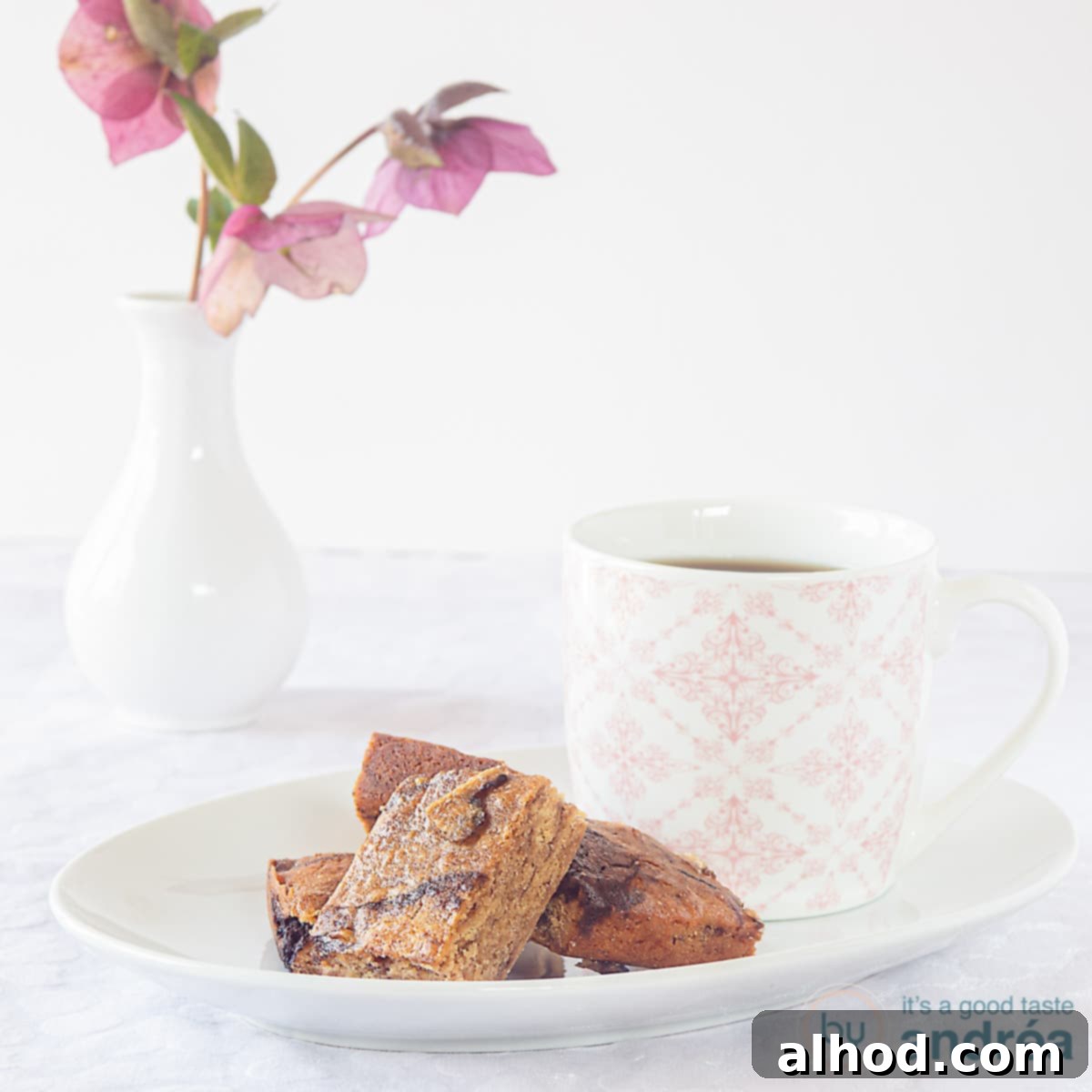Luscious Honey Blondies 3 A square photo of a white pastry plate with a few pieces of blondie with honey and chocolate. A pink and white teacup on the plate. In the background a vase with flowers.