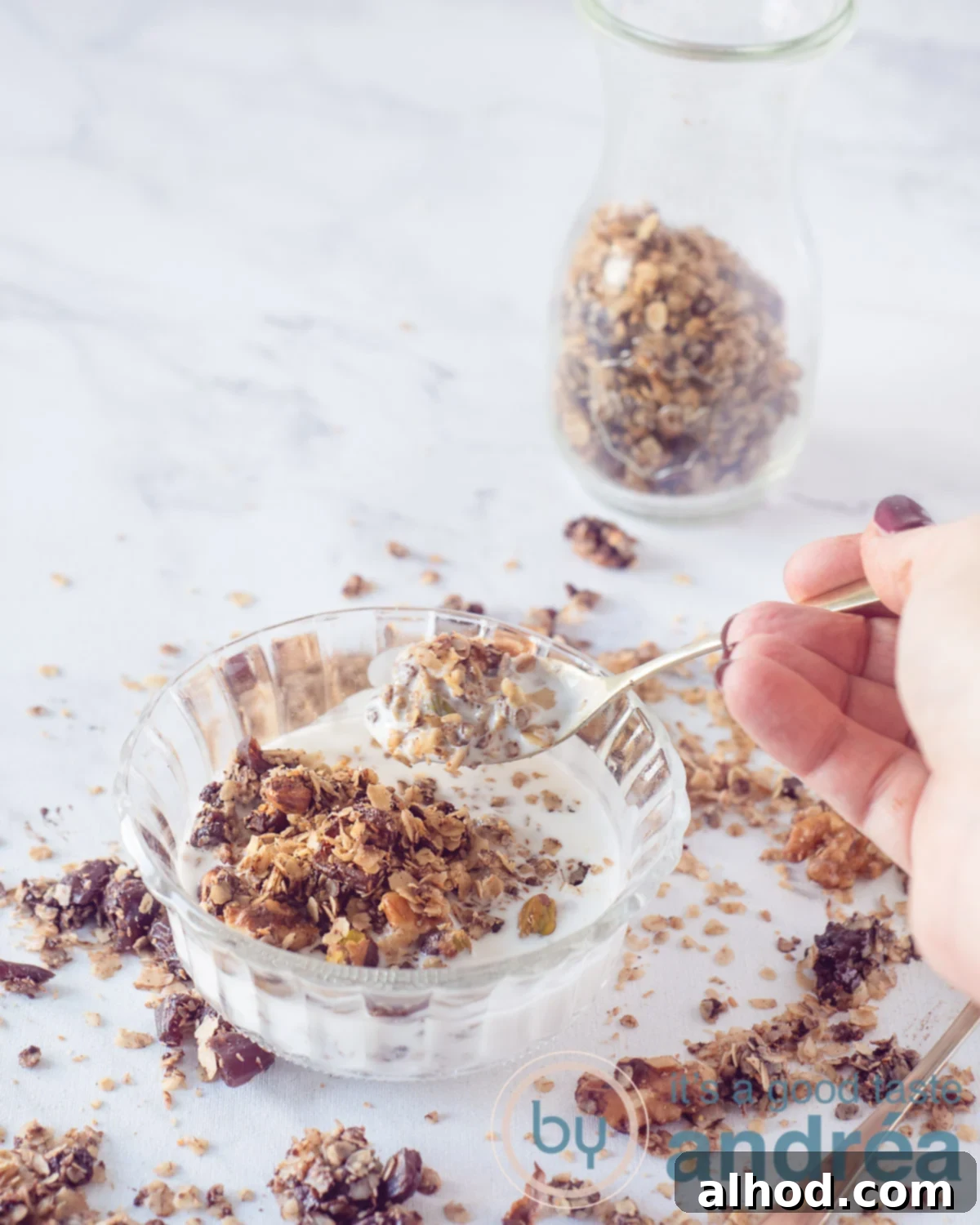 Simple Homemade Dark Chocolate Granola 2 A beautifully arranged bowl of dark chocolate nut granola with creamy yogurt. Granola pieces are artfully sprinkled around the bowl, and a golden spoon is poised for a delicious bite. In the background, a glass jar holds more freshly baked granola.