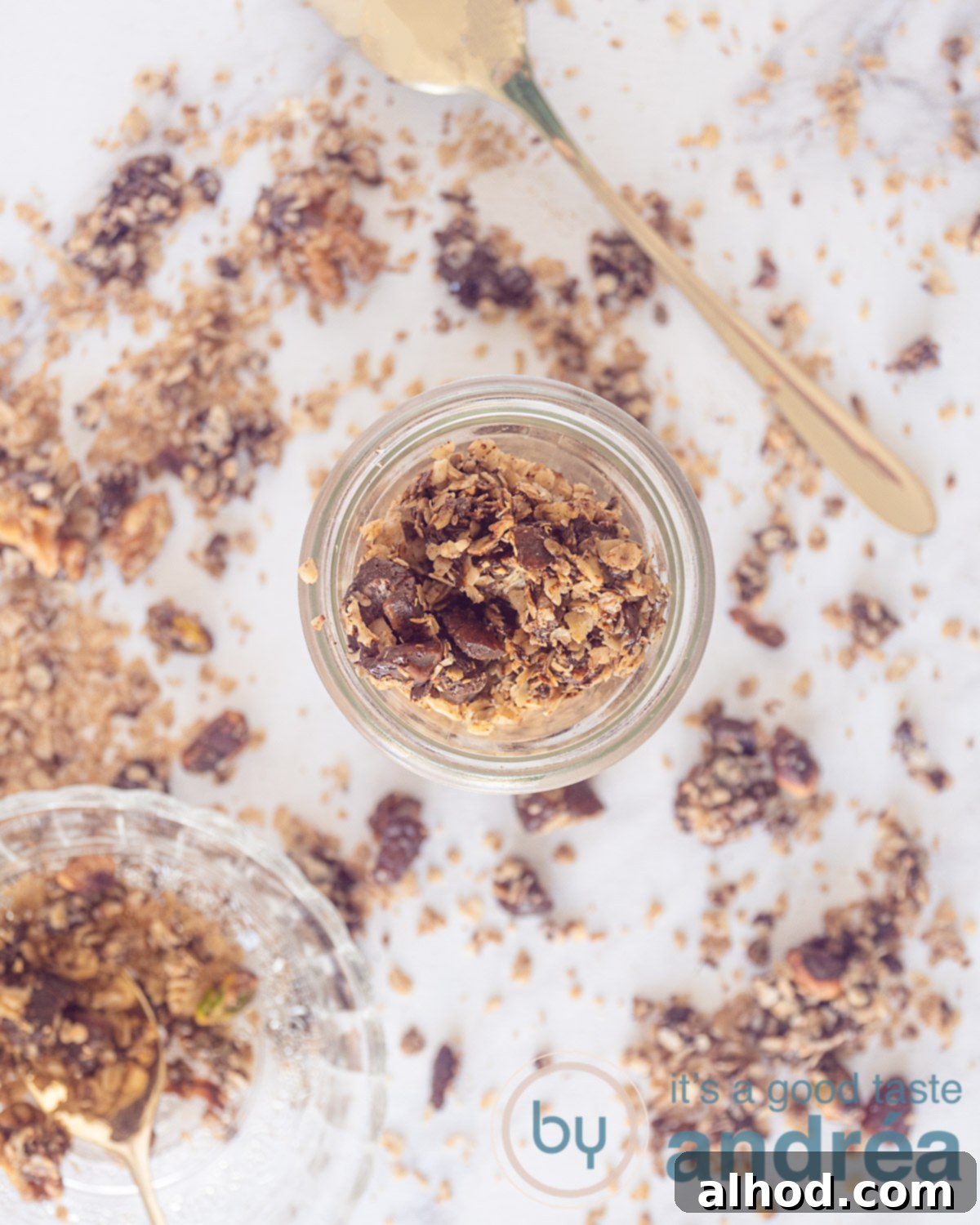 Simple Homemade Dark Chocolate Granola 3 An overhead view capturing a glass filled with tempting chocolate granola and various nuts. A bowl rests in the bottom left corner, surrounded by scattered granola pieces. A golden spoon is visible in the top right, inviting a taste.