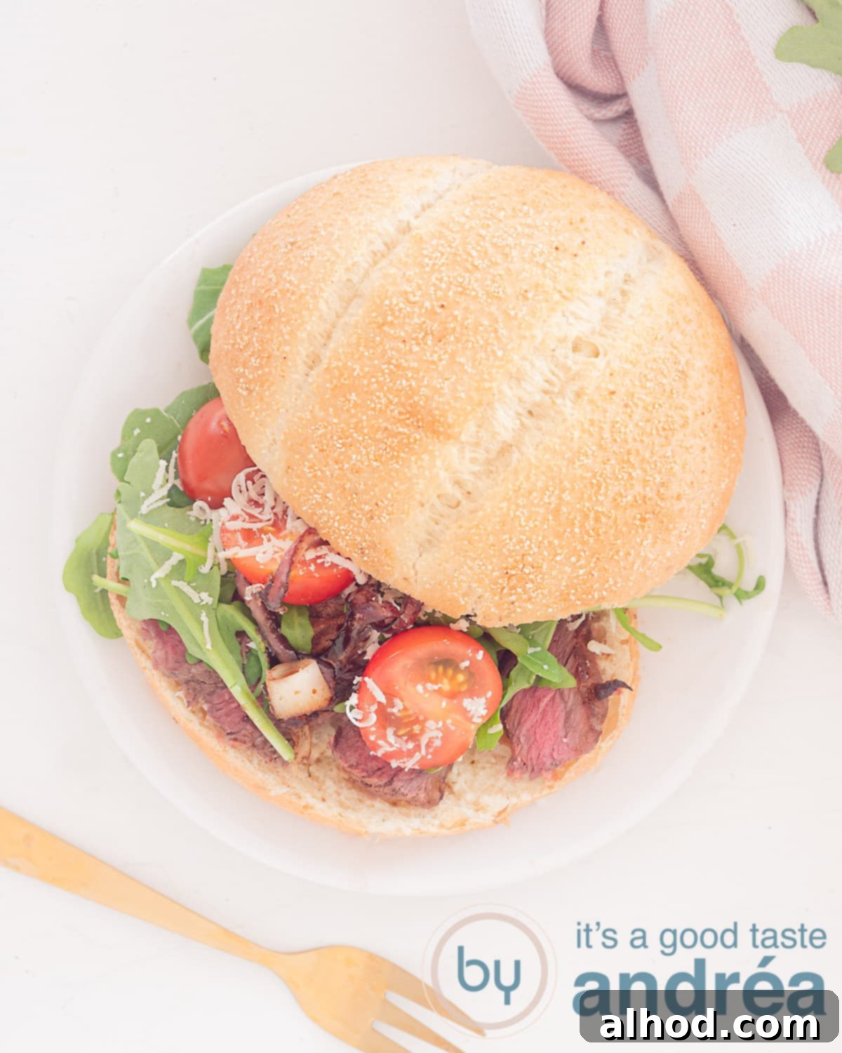A photo from above of a sandwich filled with steak, arugula, tomatoes and parmesan cheese. A fork at the bottom left. Top right a pink white tea towel.