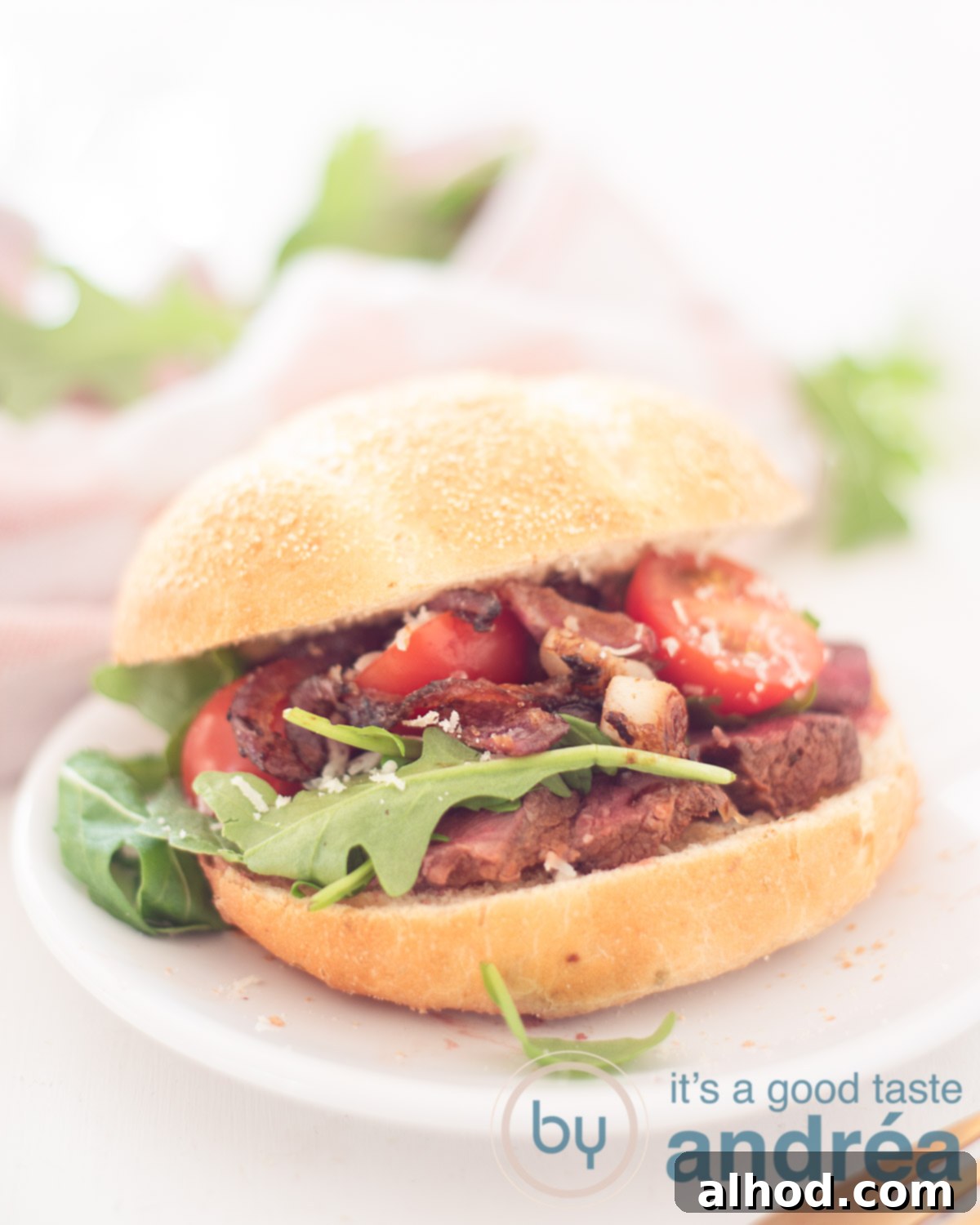 Steak Sandwich Perfection 5 Sandwich with grilled steak and caramelized onions on a white plate. In the backgroudn a pink/white cloth and some arugula.