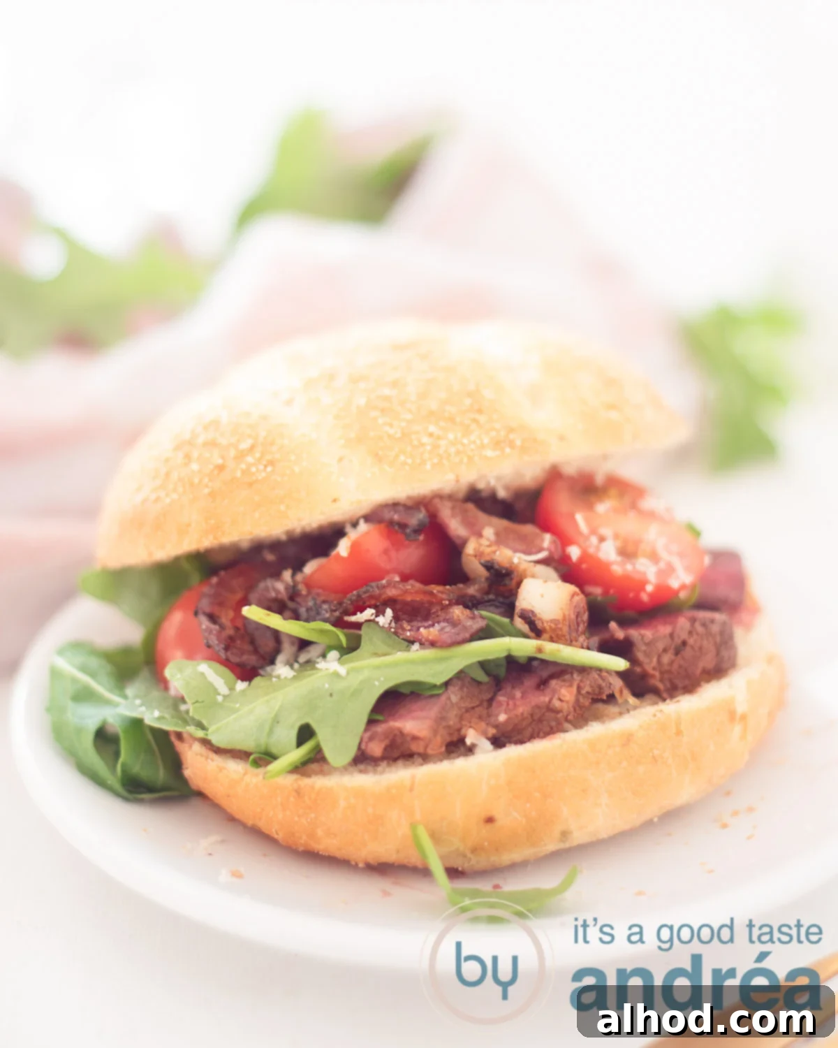 Sizzling Steak Sensation 5 Sandwich with grilled steak and caramelized onions on a white plate. In the backgroudn a pink/white cloth and some arugula.