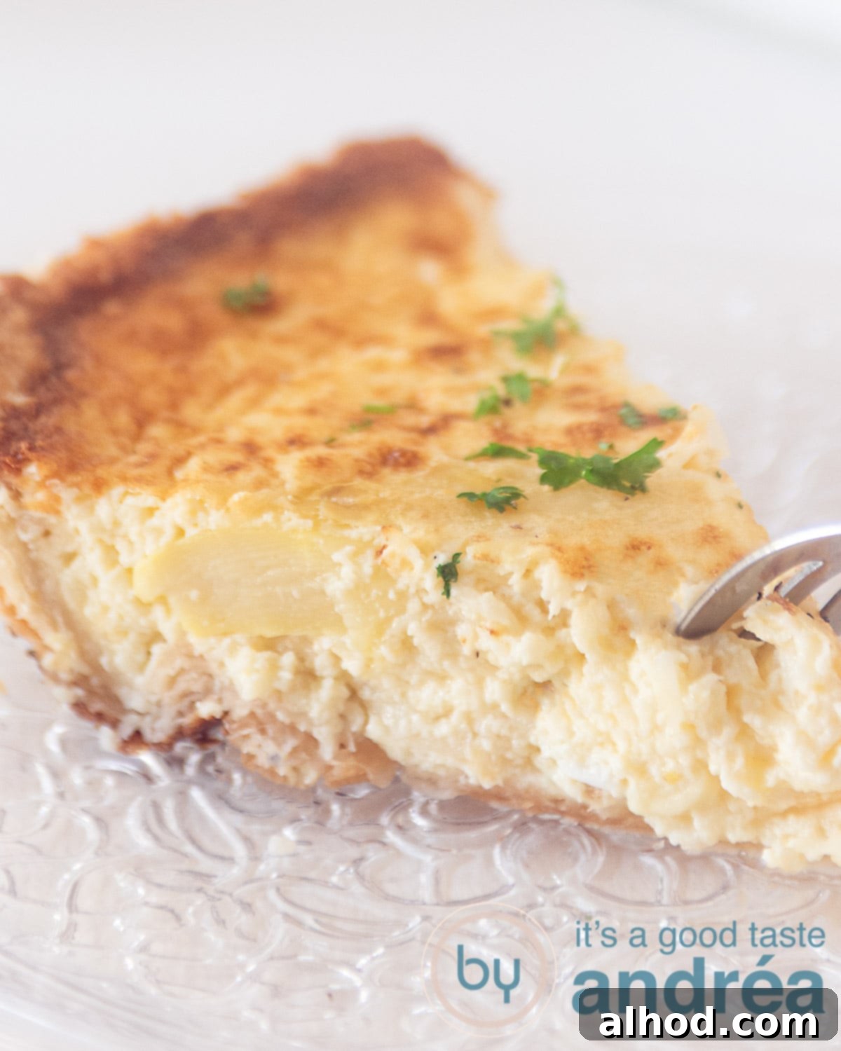 Vegetarian White Asparagus Quiche 2 A slice of a quiche with asparagus and cheeses on a glass plate with a fork taking a bite. The background is white.