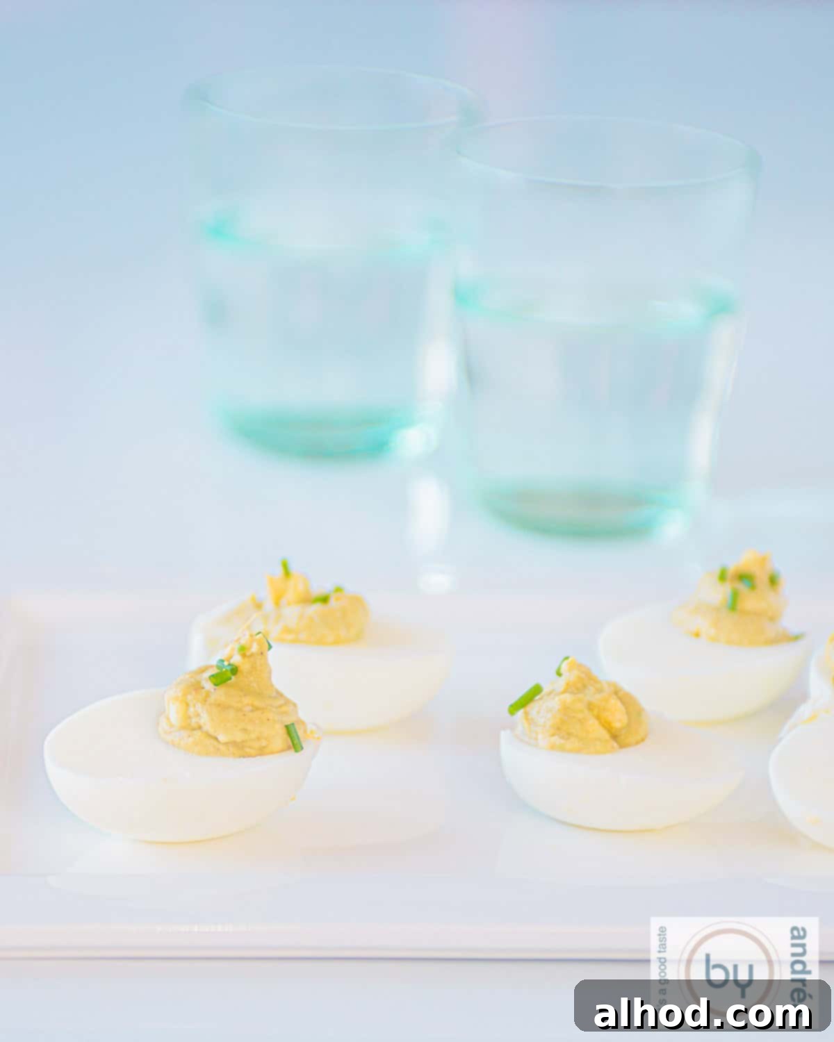 Velvet Avocado Deviled Eggs 2 A flat white plate filled with perfectly piped avocado deviled eggs, garnished with fresh chives. Two elegant blue glasses are blurred in the background, hinting at a sophisticated setting.