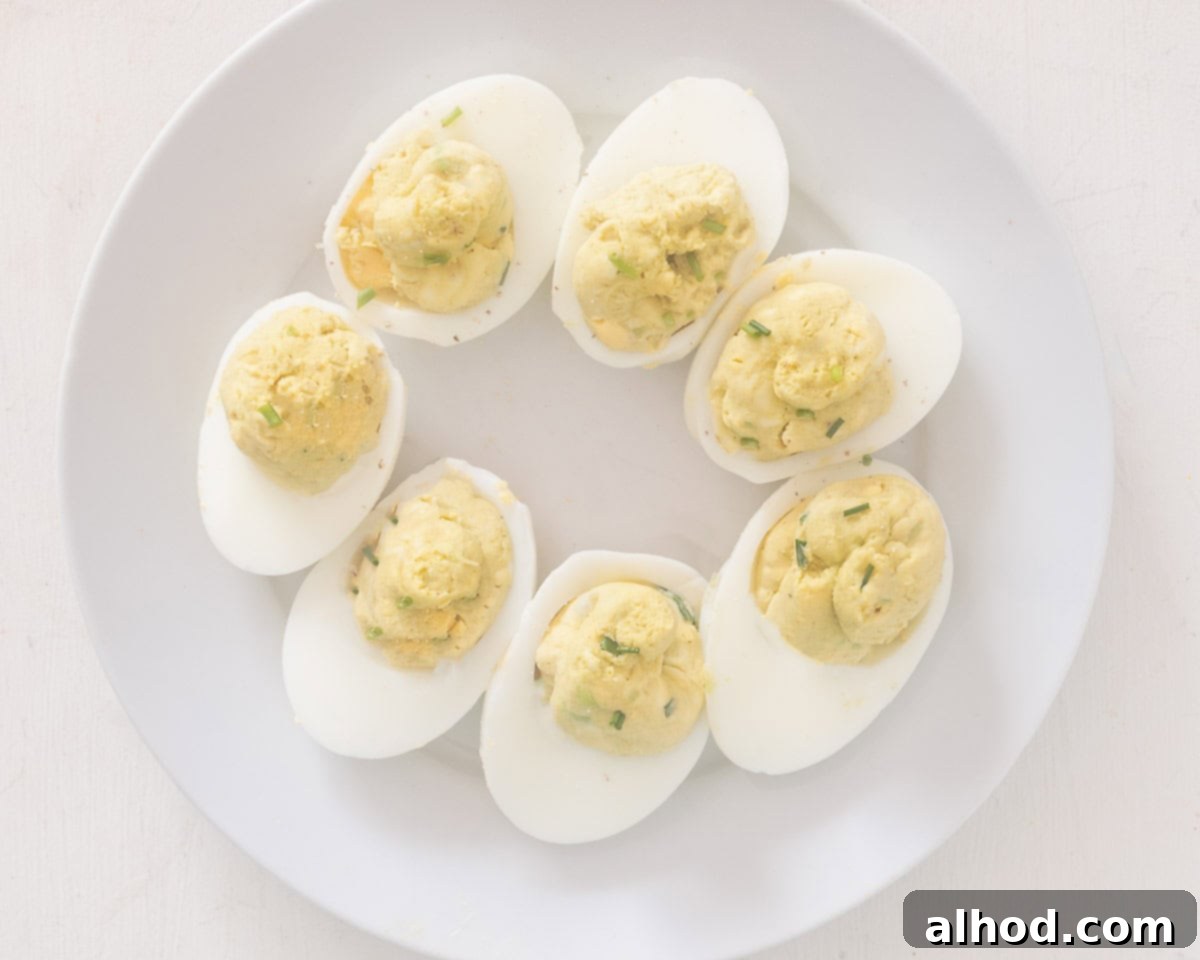 Velvet Avocado Deviled Eggs 7 A pristine white plate showcasing several finished avocado deviled eggs, artfully arranged in a circular pattern and generously sprinkled with finely chopped fresh chives for a pop of color and flavor.