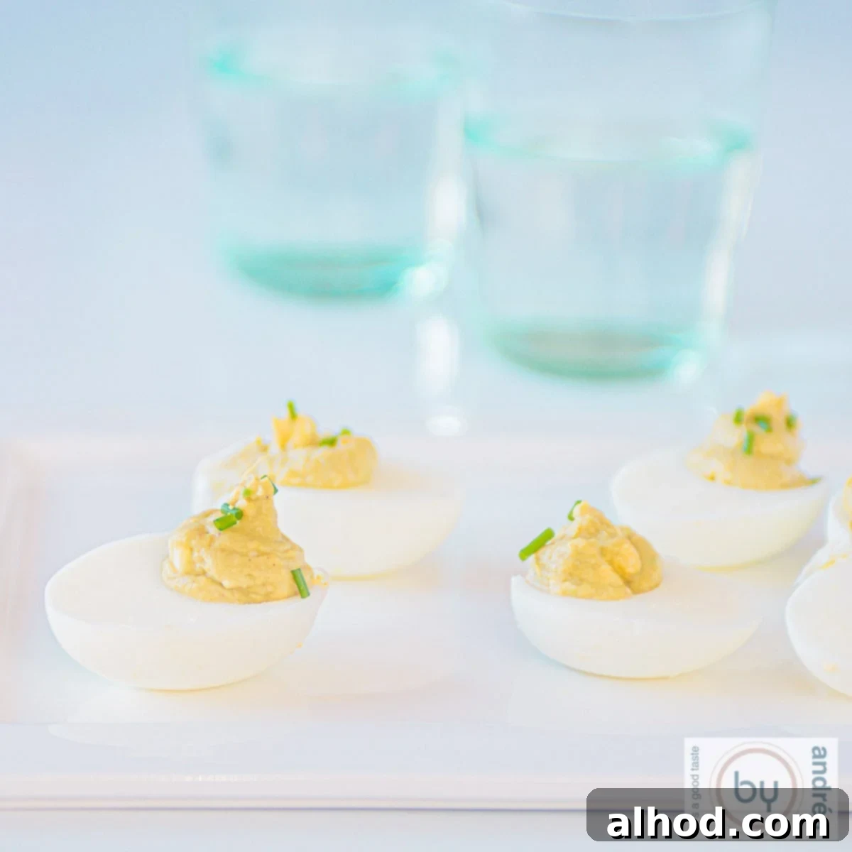 Velvet Avocado Deviled Eggs 9 A square picture showing a white plate with a dozen avocado curry deviled eggs, elegantly arranged and garnished with fresh herbs. Two blue glasses are visible in the soft background.