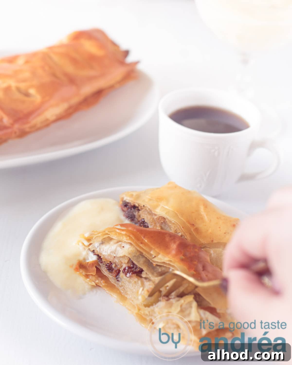 A beautifully baked German Apple Strudel on a white plate, ready to be served. A hand reaches for a slice, with coffee and other pastries softly blurred in the background, hinting at a cozy tea time.