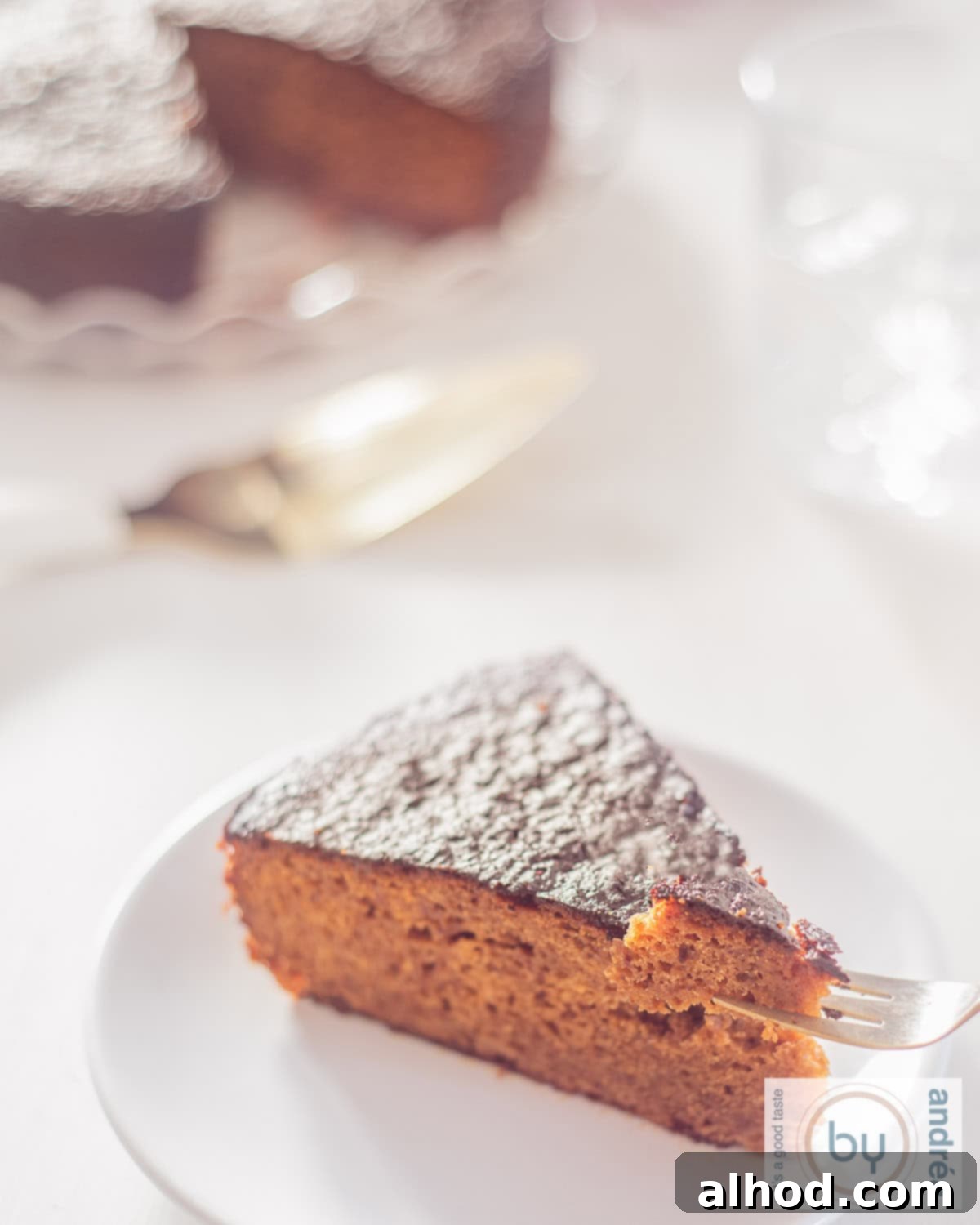 Melt In Your Mouth Sticky Toffee Cake 3 A photo of a piece of sticky date cake on a white plate. A fork pricks a bite off. More sticky toffee pudding in the background