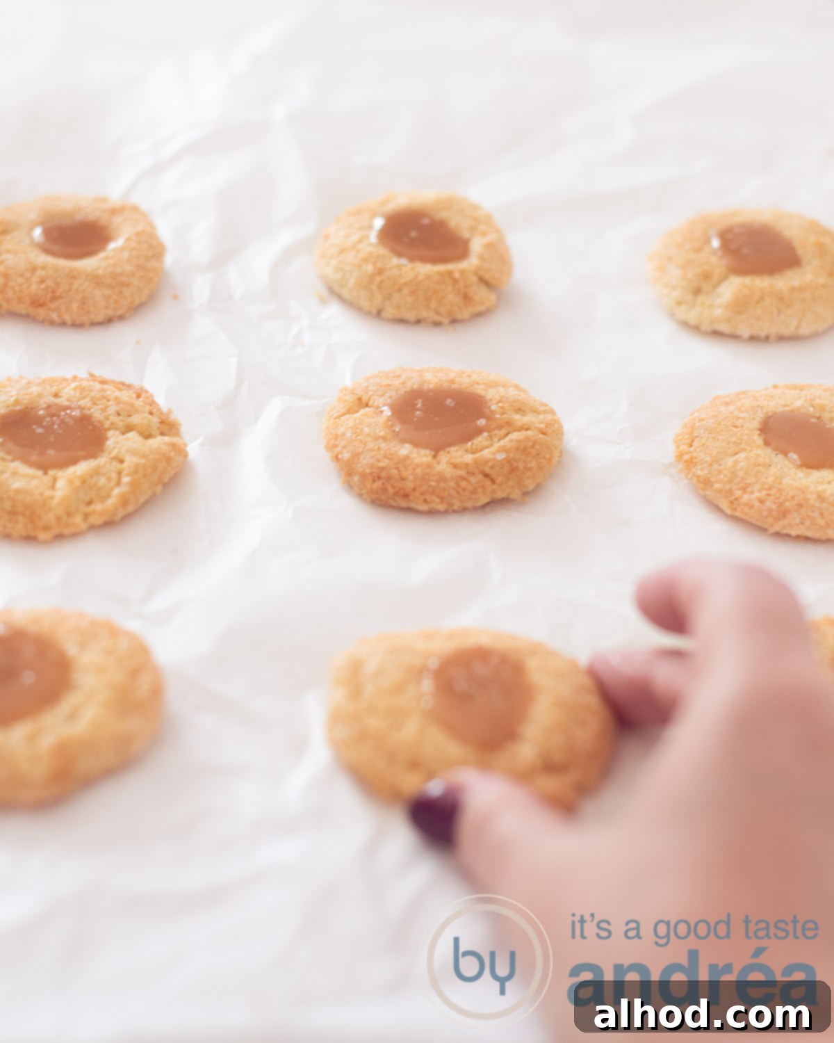 Decadent Coconut Caramel Cookies 5 8 coconut cookies filled with caramel and seasalt on a white baking paper. A hand is taking a cookie.