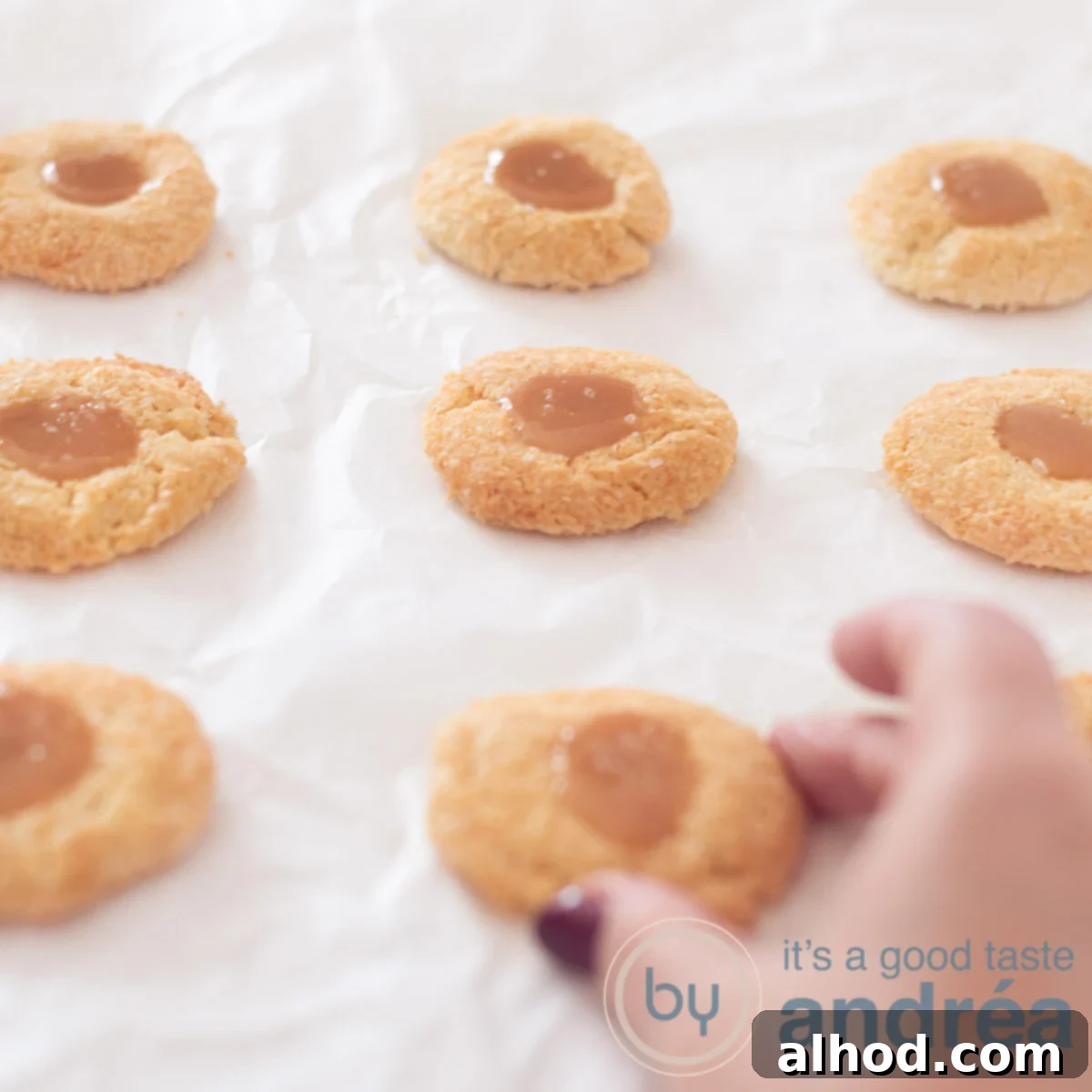 Decadent Coconut Caramel Cookies 6 a square picture with eight coconut caramel cookies on a white baking paper. A hand is grabbing the middle one.