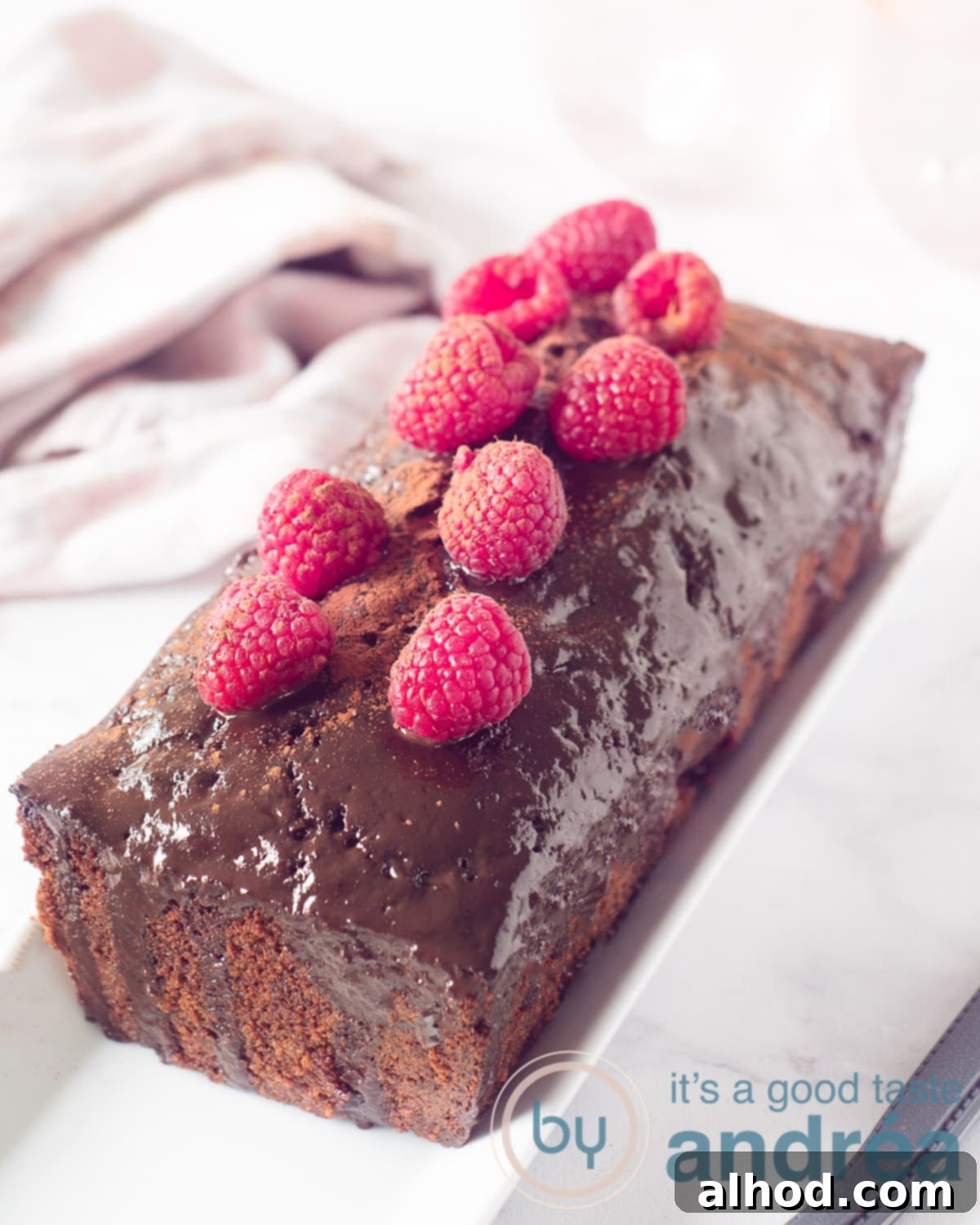 Chocolate Raspberry Loaf Delight 2 A height picture with a raspberry chocolate loaf cake topped with chocolate ganache and fresh strawberries. A white pink cloth in the back.