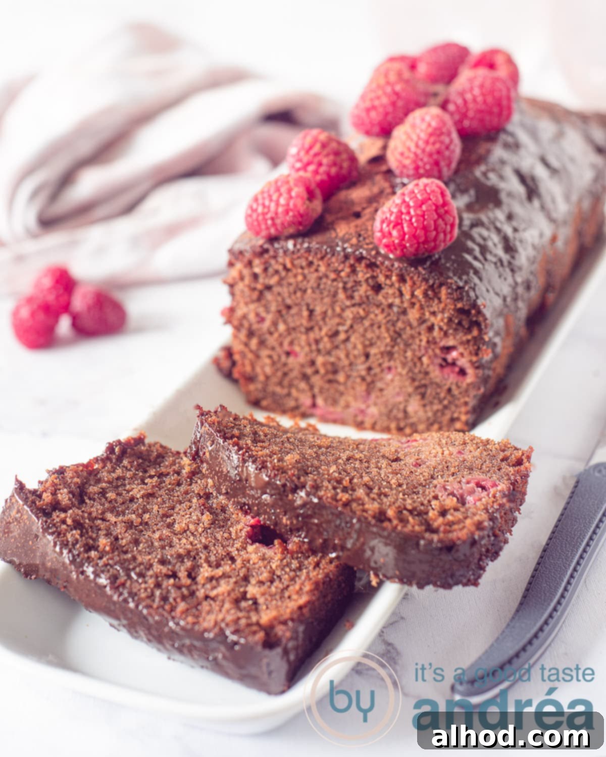 Chocolate Raspberry Loaf Delight 7 A loaf of chocolate raspberry cake, topped with ganache and raspberries. Some raspberries and a pink white cloth in the background.