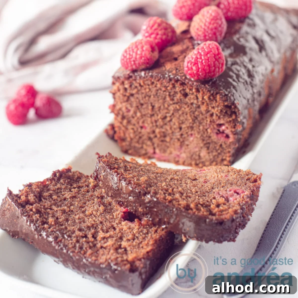 A square photo with a chocolate cake with raspberries. Two slices in front. In the back more raspberries and a white pink cloth.