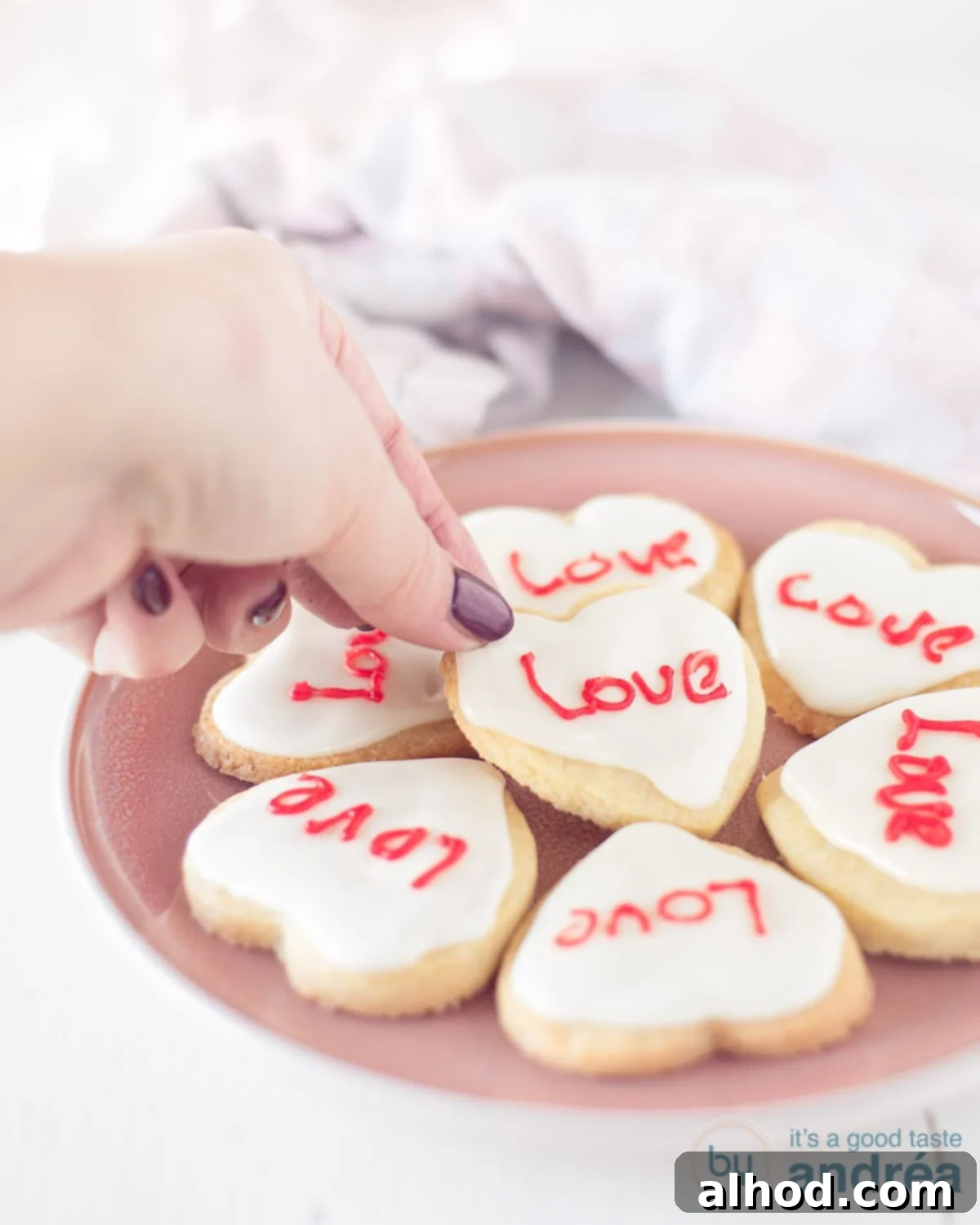 Sweetheart Cookies 3 A pink plate with 7 heart cookies with love written on them. A hand grabs a cookie.