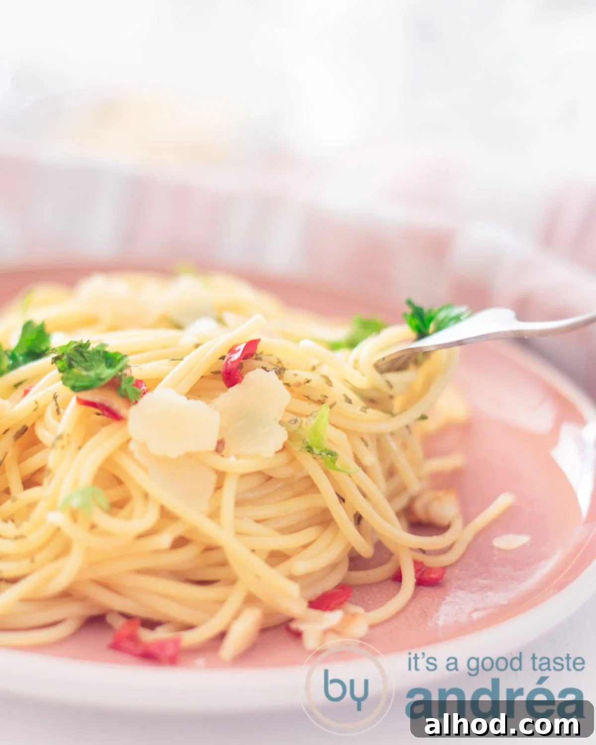 Rustic Chili Garlic Pasta 5 Part of a pink plate with a mountain of spaghetti with peppers, parsley, parmesan cheese, garlic and olive oil. A fork sticks into the spaghetti. A white background