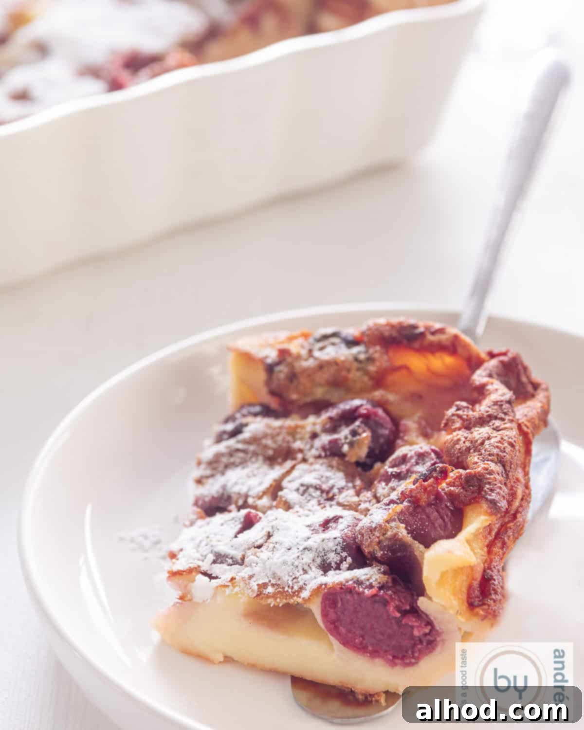 Rustic French Cherry Clafoutis 2 A perfectly portioned slice of golden-brown Cherry Clafoutis on a pristine white plate, with more clafoutis visible in a rustic ceramic bowl in the soft-focus background.