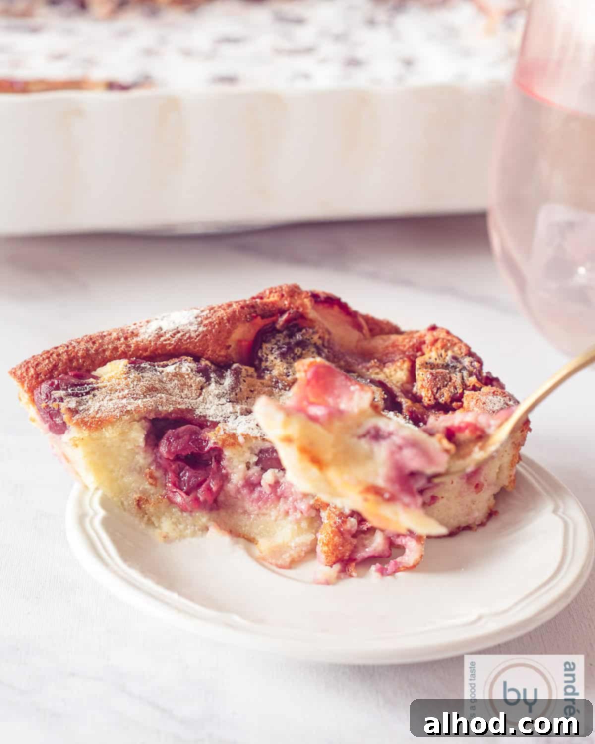 Rustic French Cherry Clafoutis 3 An overhead shot showcasing a golden-brown cherry clafoutis in an oven dish at the back, with a single serving on a white plate in the foreground, a fork having just taken a delicious bite.