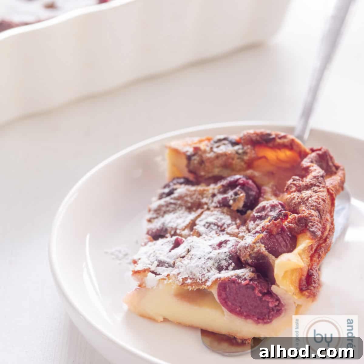 Rustic French Cherry Clafoutis 4 A square picture of a white plate with a slice of Cherry Clafoutis, showcasing its creamy texture and juicy cherries.