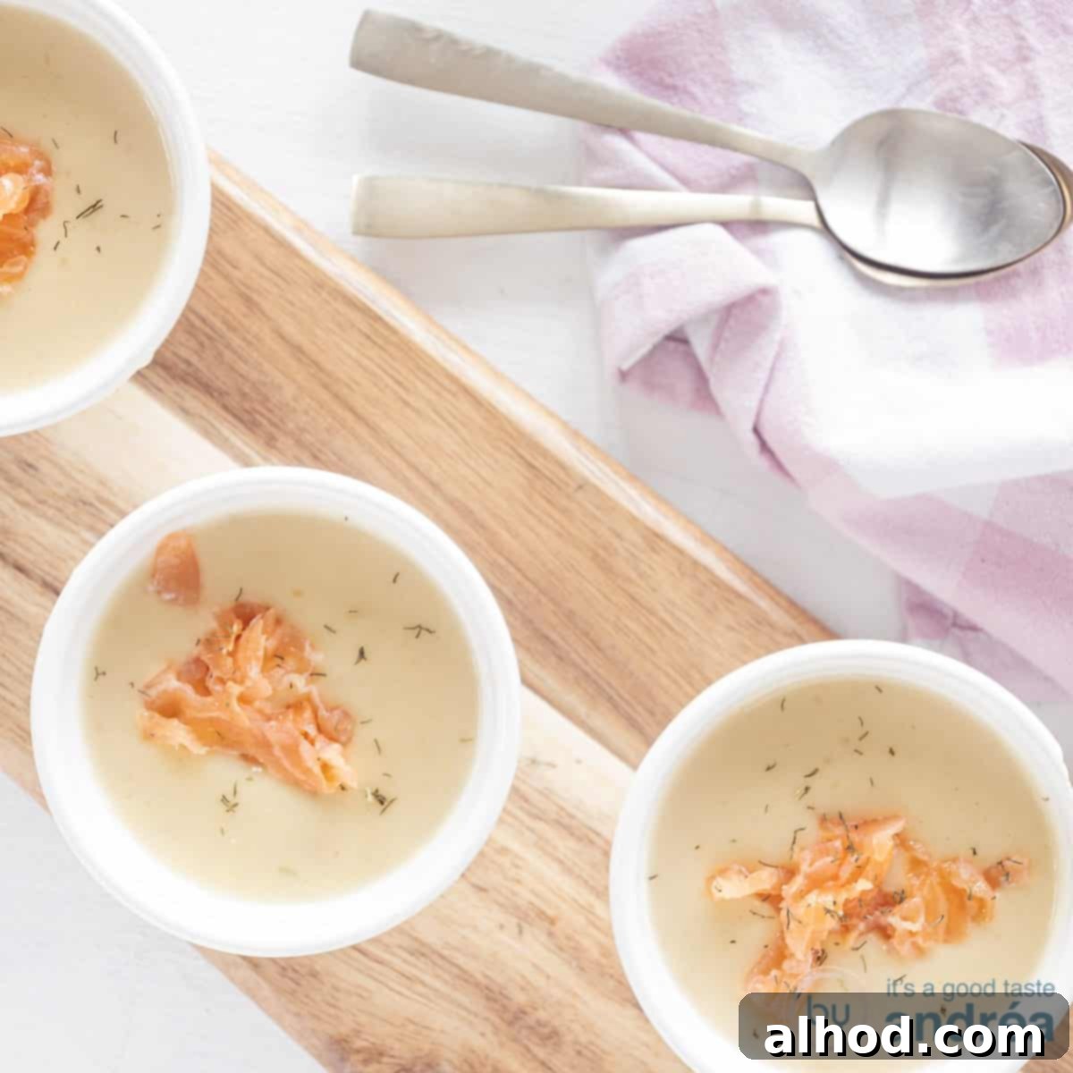 A square photo from above with three white cups filled with thickened queen soup with a garnish of smoked salmon and dill. At the top right a pink white tea towel and two gold spoons.