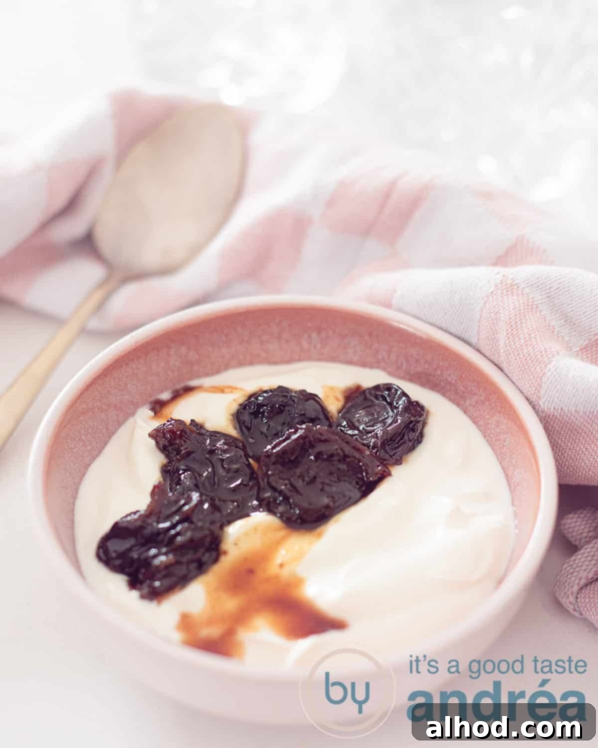 A pink bowl filled with creamy Greek yogurt, rich plum compote, and a drizzle of sweet sauce. In the background, a soft pink and white cloth and a golden spoon hint at a cozy, inviting setting.