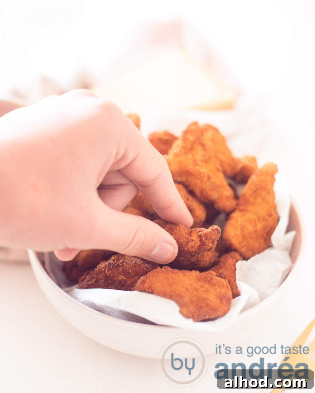 Parmesan Crusted Chicken Nuggets 2 A hand reaches for a golden-brown Parmesan Chicken Nugget from a pink bowl, set against a clean white background.