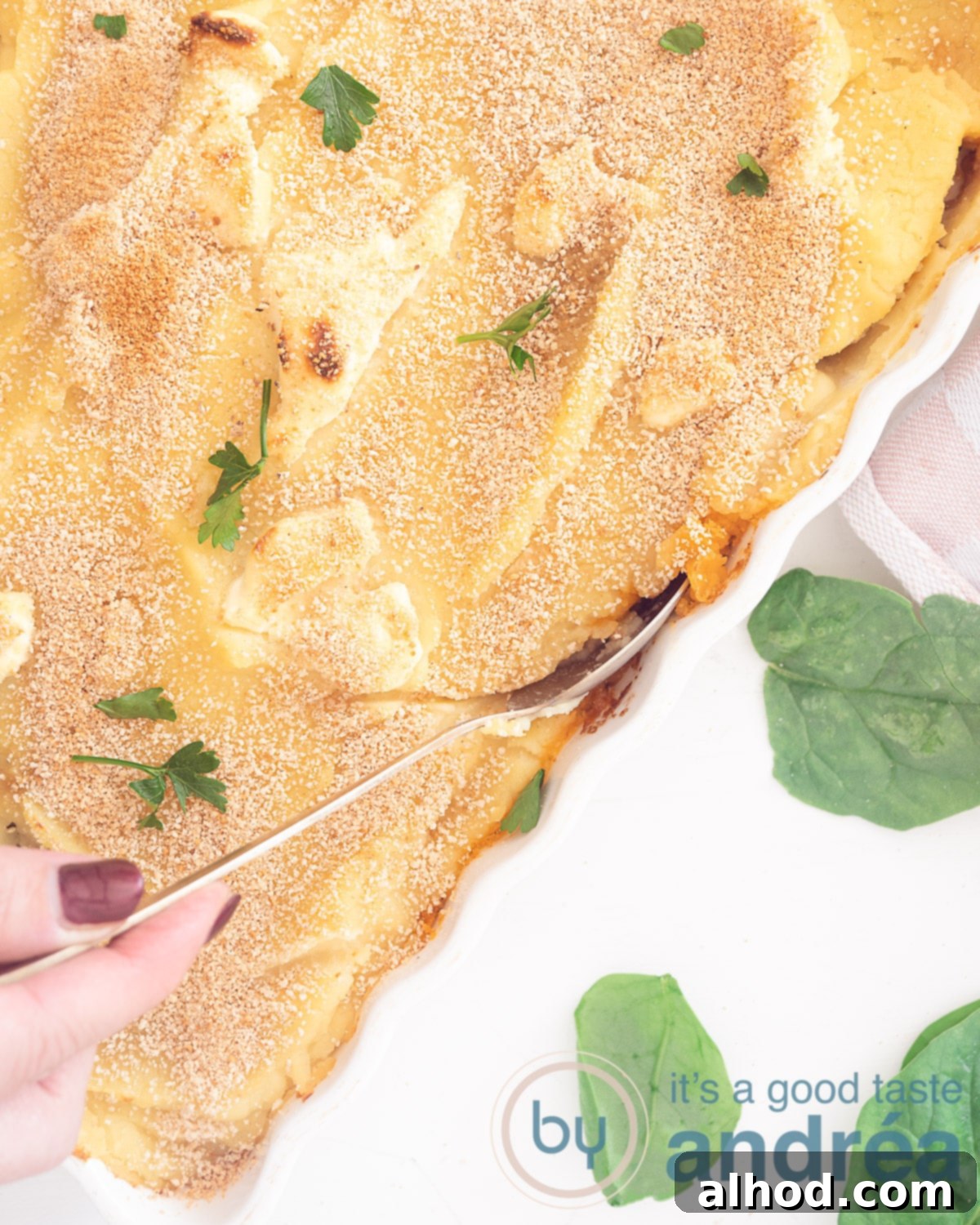 Comforting Beef, Potato and Spinach Bake 3 A top-down shot of a white baking dish filled with a savory gratin. A hand is scooping a generous portion with a spoon. Fresh spinach leaves are visible in the bottom left corner, emphasizing the healthy ingredients.