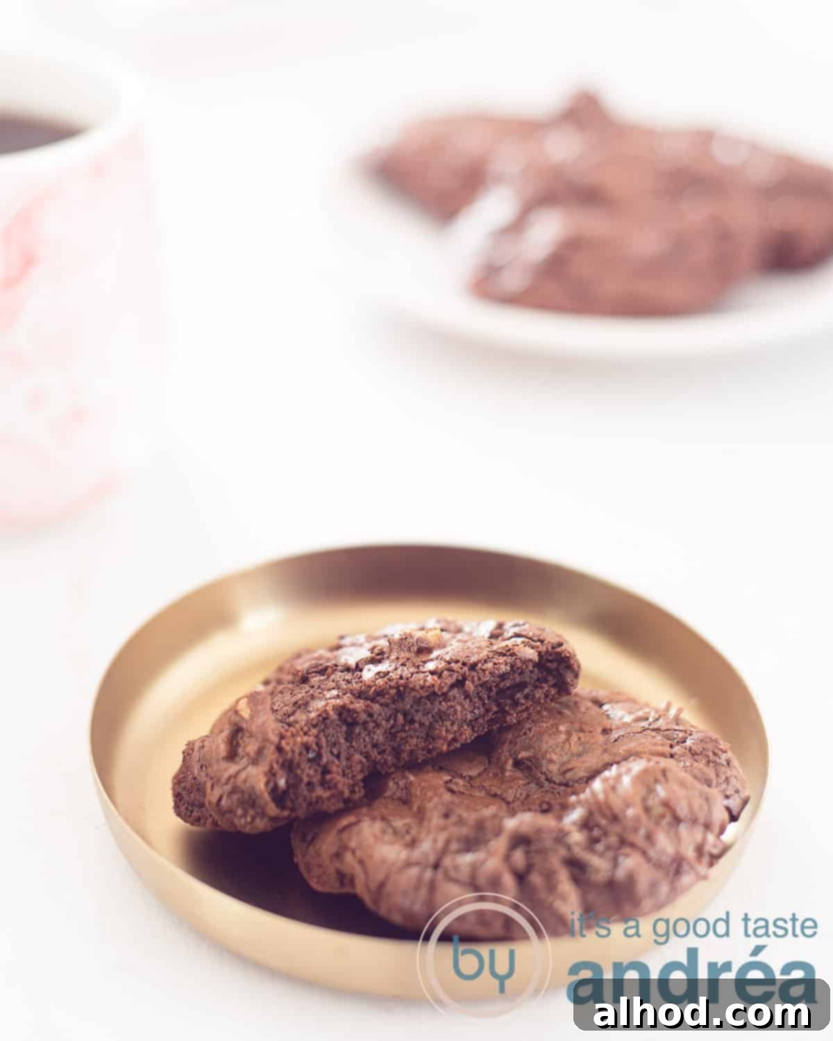 Fudgy Brownie Walnut Cookies 2 A golden bowl with a brownie cookie and half a cookie on top, so that you can see the airy inside. On a white background. In the background a cup of coffee and a plate with more cookies.