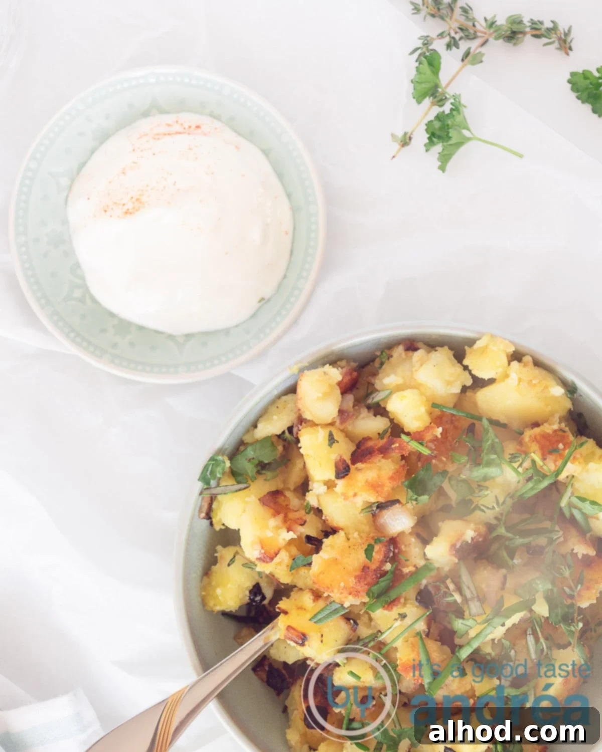 A top-down photo of a bowl of pan-fried potatoes with herbs and a blue bowl of garlic yogurt sauce on a white background, with fresh herbs scattered around.
