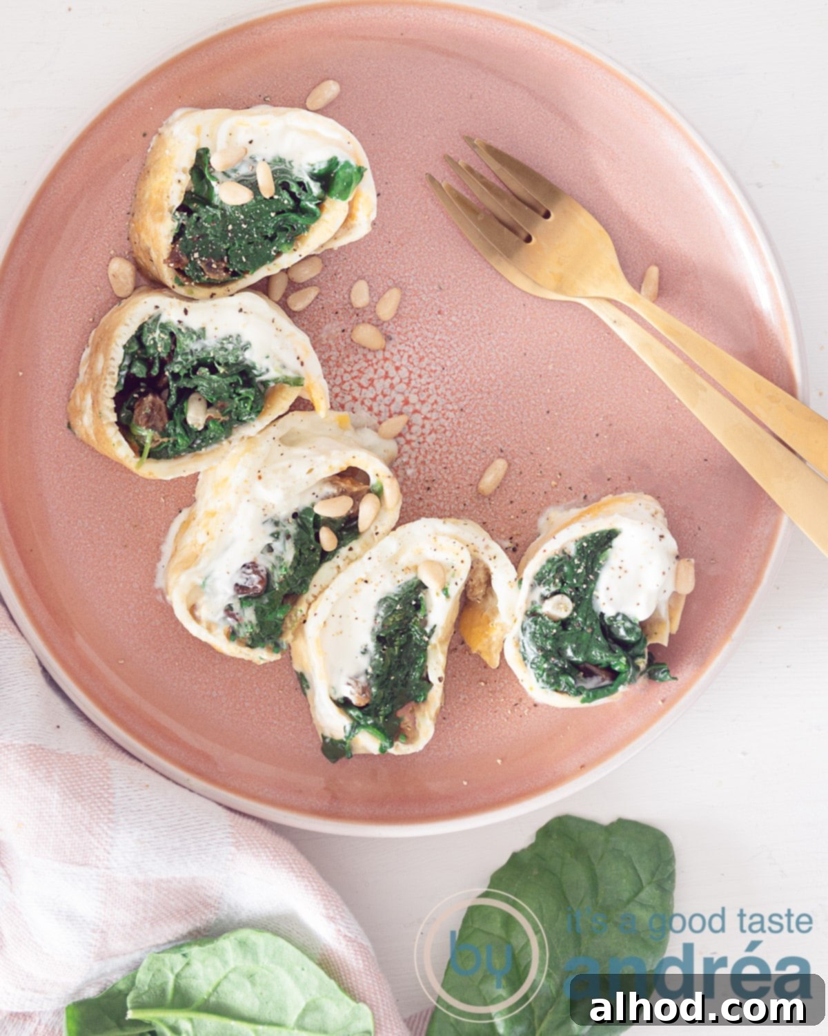 Savory Spinach and Pine Nut Omelet Rolls 3 A photo from above with a pink plate at the top with a half moon of spinach sour cream wraps. Next to it two golden forks. Spinach leaves at the bottom