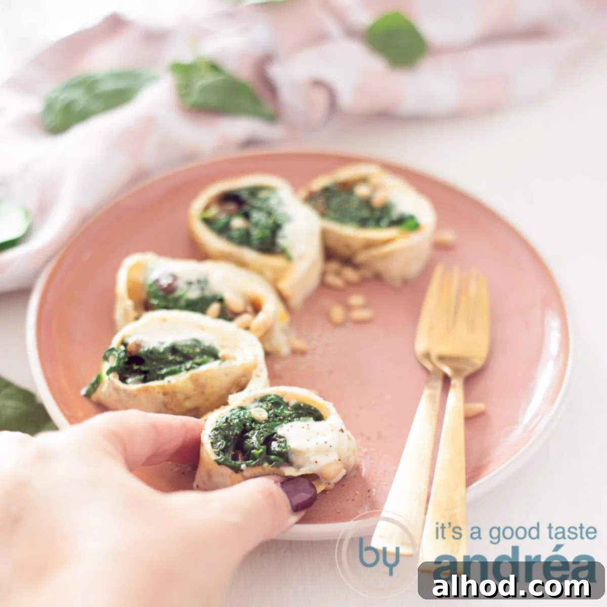 Savory Spinach and Pine Nut Omelet Rolls 4 A square photo with a pink plate in the middle on the right side two gold forks. Around it vegetarian wrap snacks with spinach. A hand grabs a bite. At the top a pink white tea towel with spinach leaves.