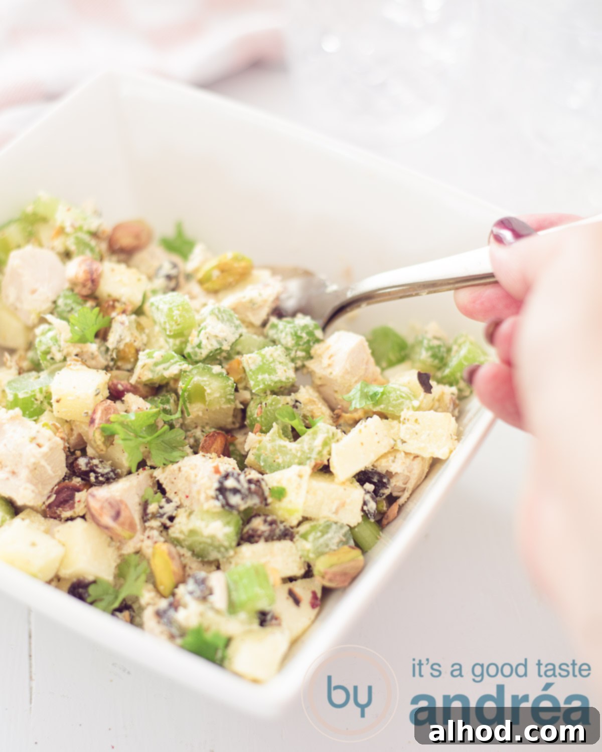 An apple chicken waldorf salad in a white square bowl. A hand is taken a spoonfull out. A white background