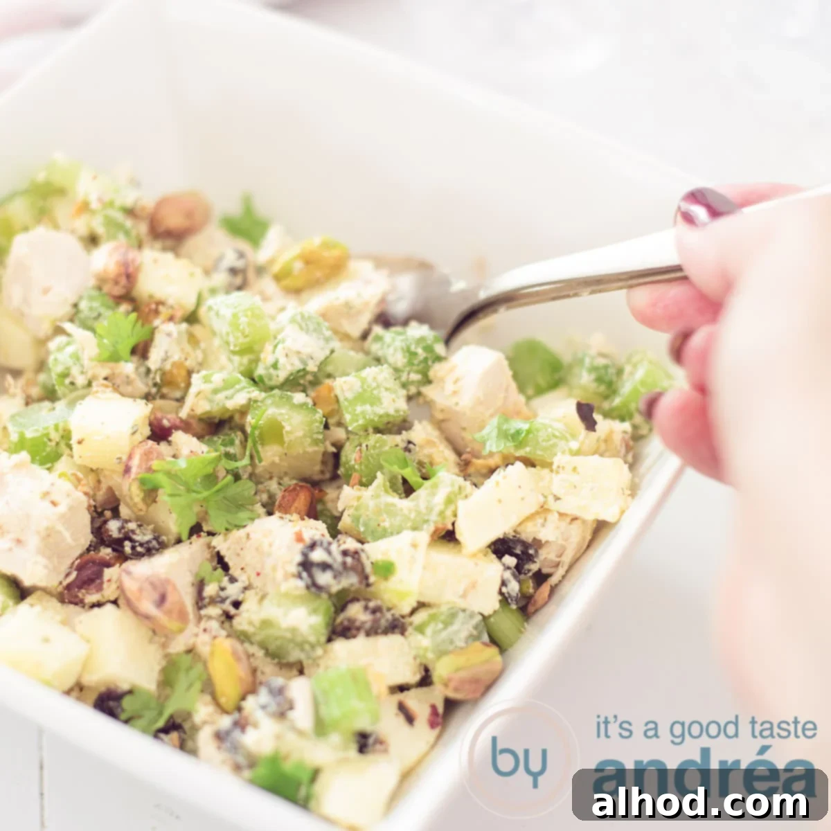 A square photo of a square white bowl filled with Waldorf salad. A hand is holding a spoon grabbing some salad. On a white background