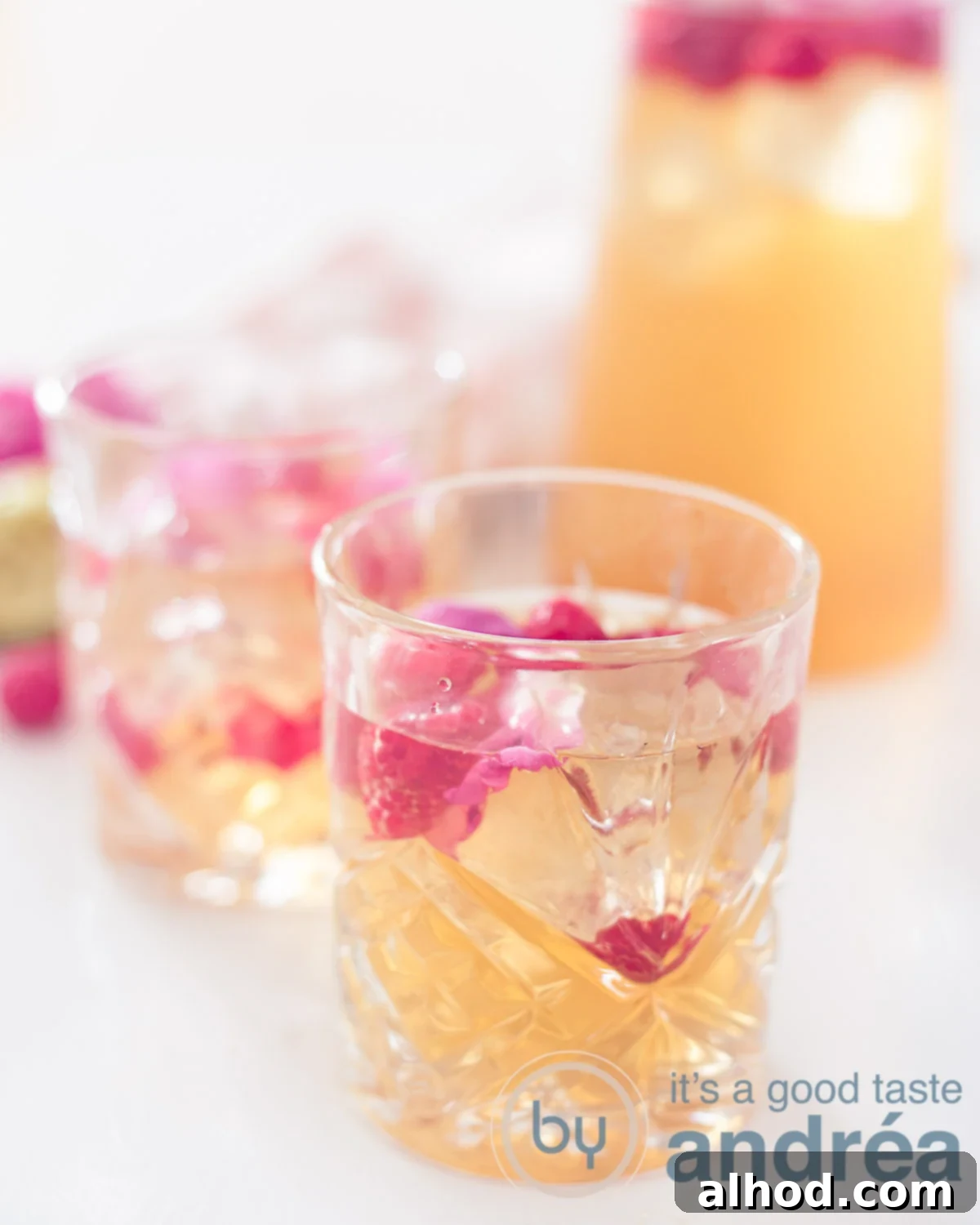 Aromatic Rose & Zesty Lime Iced Tea 2 A jar of Lime rose ice tea with raspberries and two glasses filled with ice tea on a white background, highlighting its refreshing appeal.